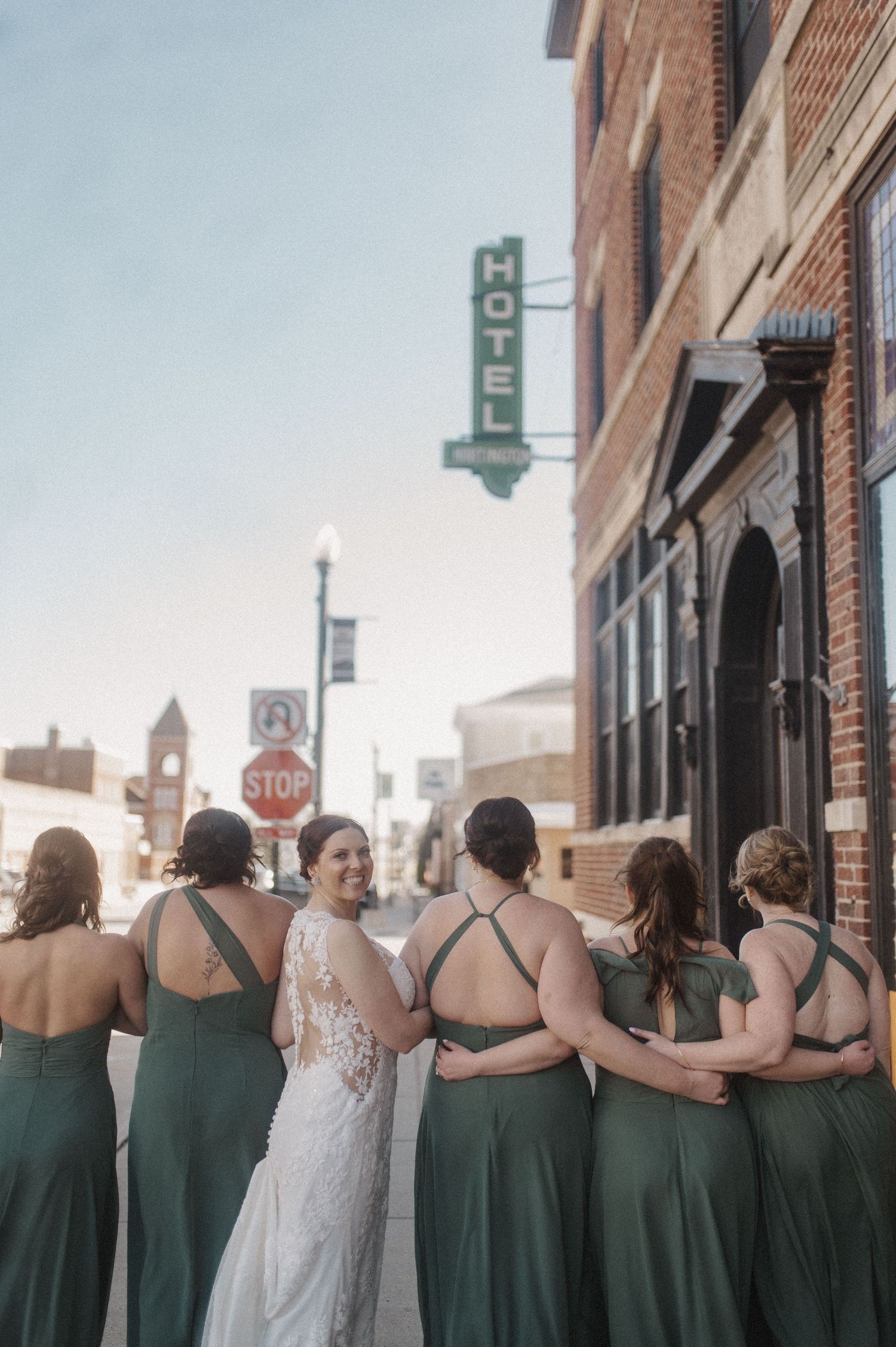 Bridal party in emerald dresses with arms around each other outside brick building.