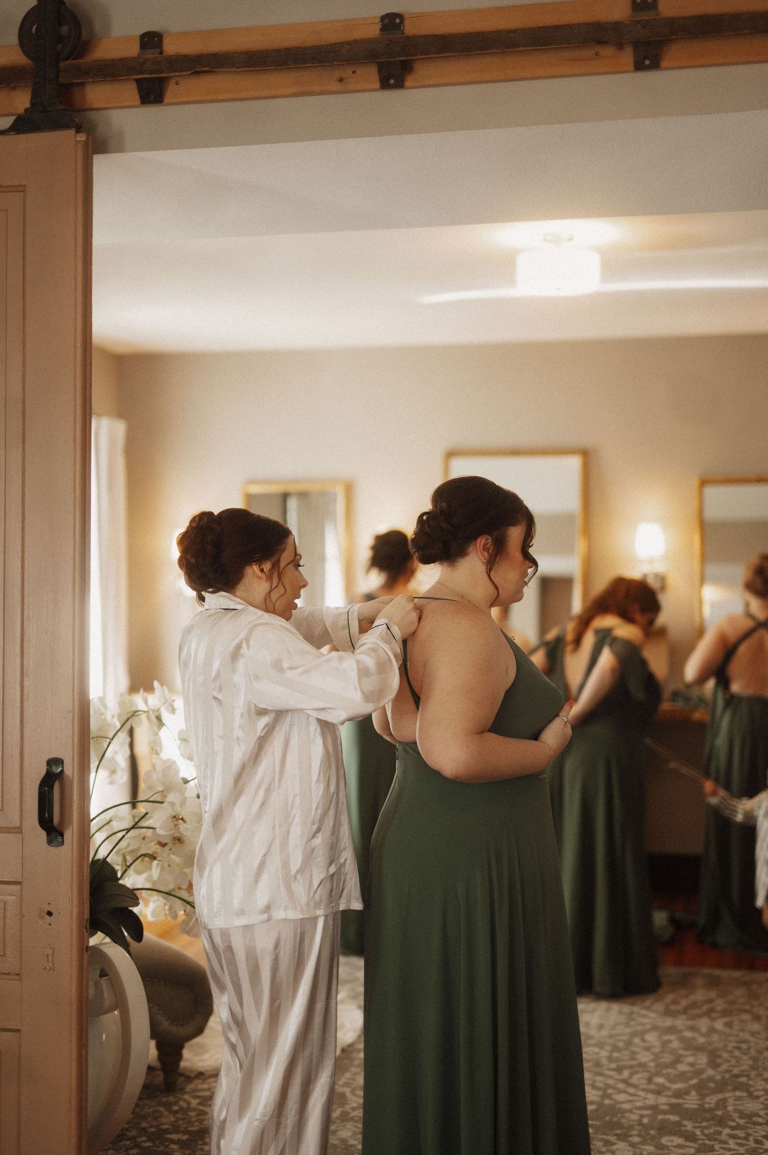 Bridesmaids in forest green dresses help with wedding preparations in a bright indoor setting.