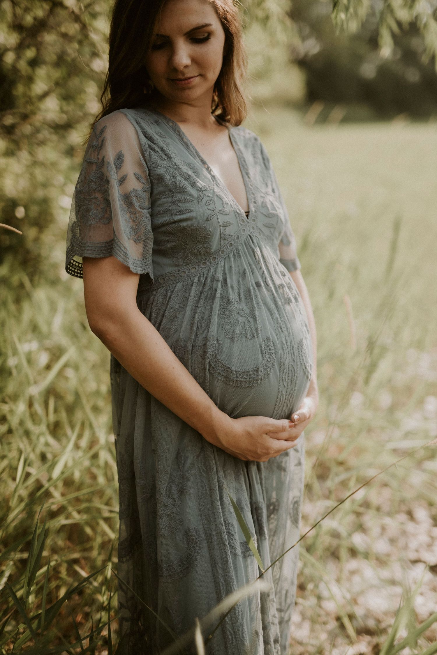 A person in a gray dress stands in a sunlit meadow showing their pregnant belly in a maternity photoshoot series.