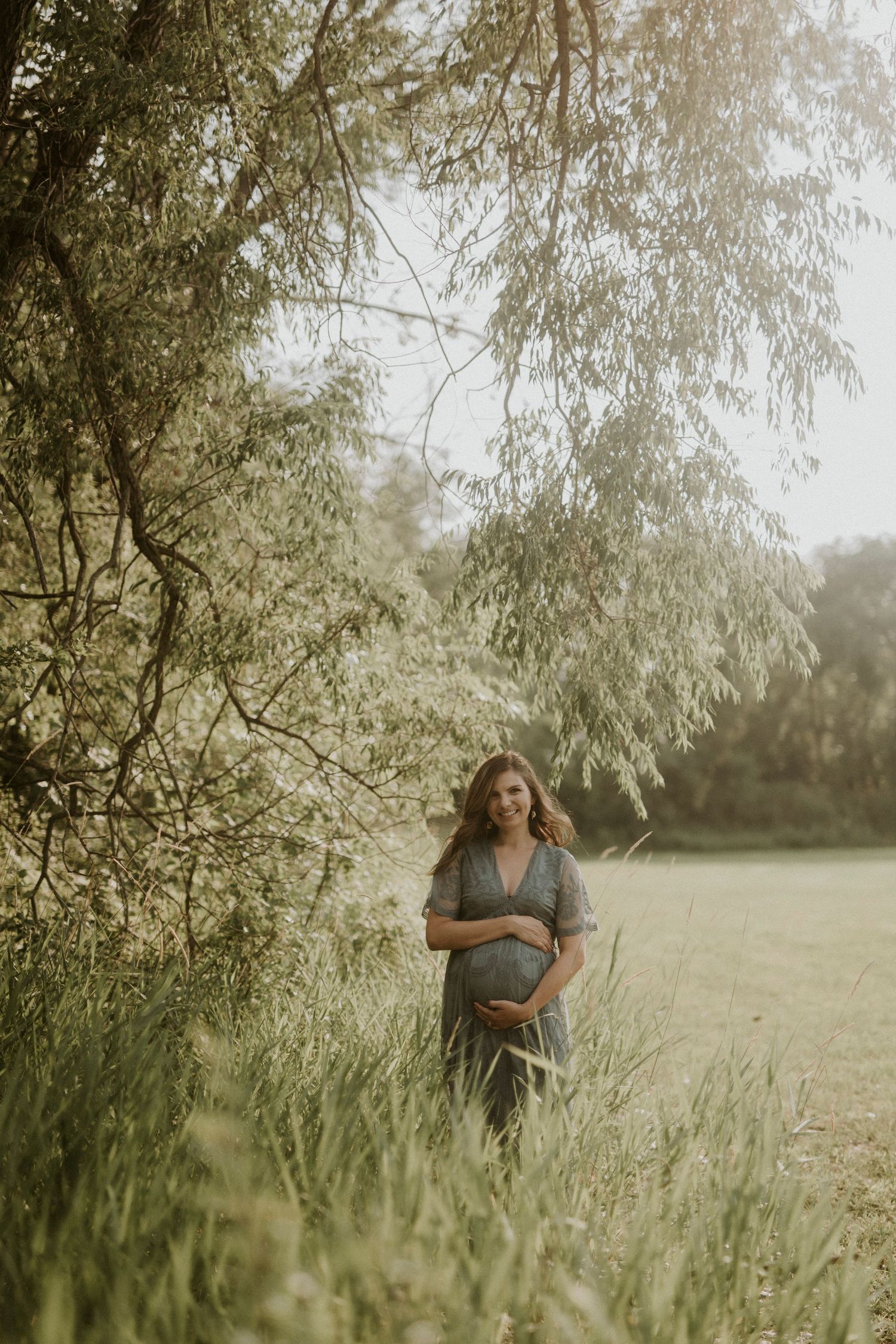 A maternity photoshoot in a serene meadow with willow trees and soft natural lighting creates a dreamy atmosphere.