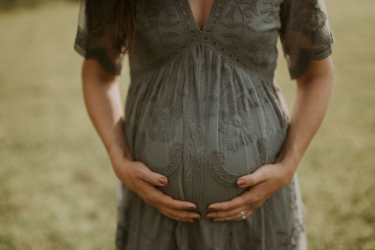 A pregnant woman in a gray dress holds her belly while standing in a sunlit outdoor setting.