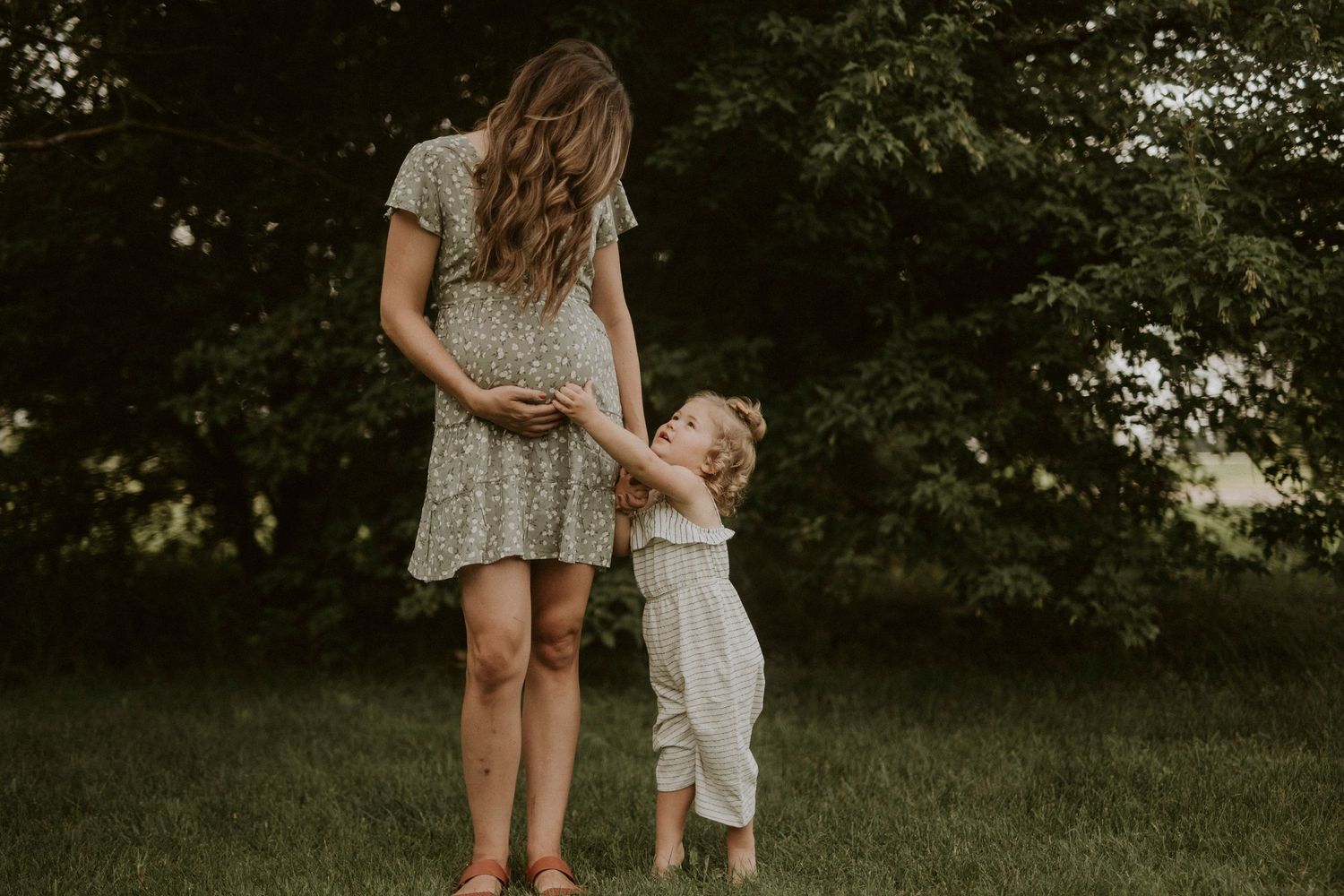 Mother and child in matching floral dresses stand together in a sunlit garden setting.