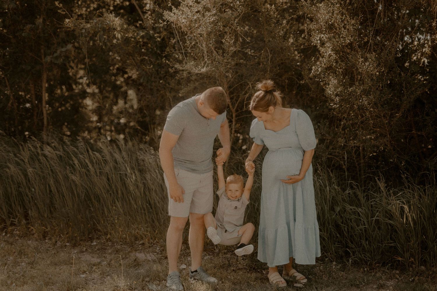 A couple and their child walk hand in hand through a wooded area at sunset creating a silhouette against warm light.