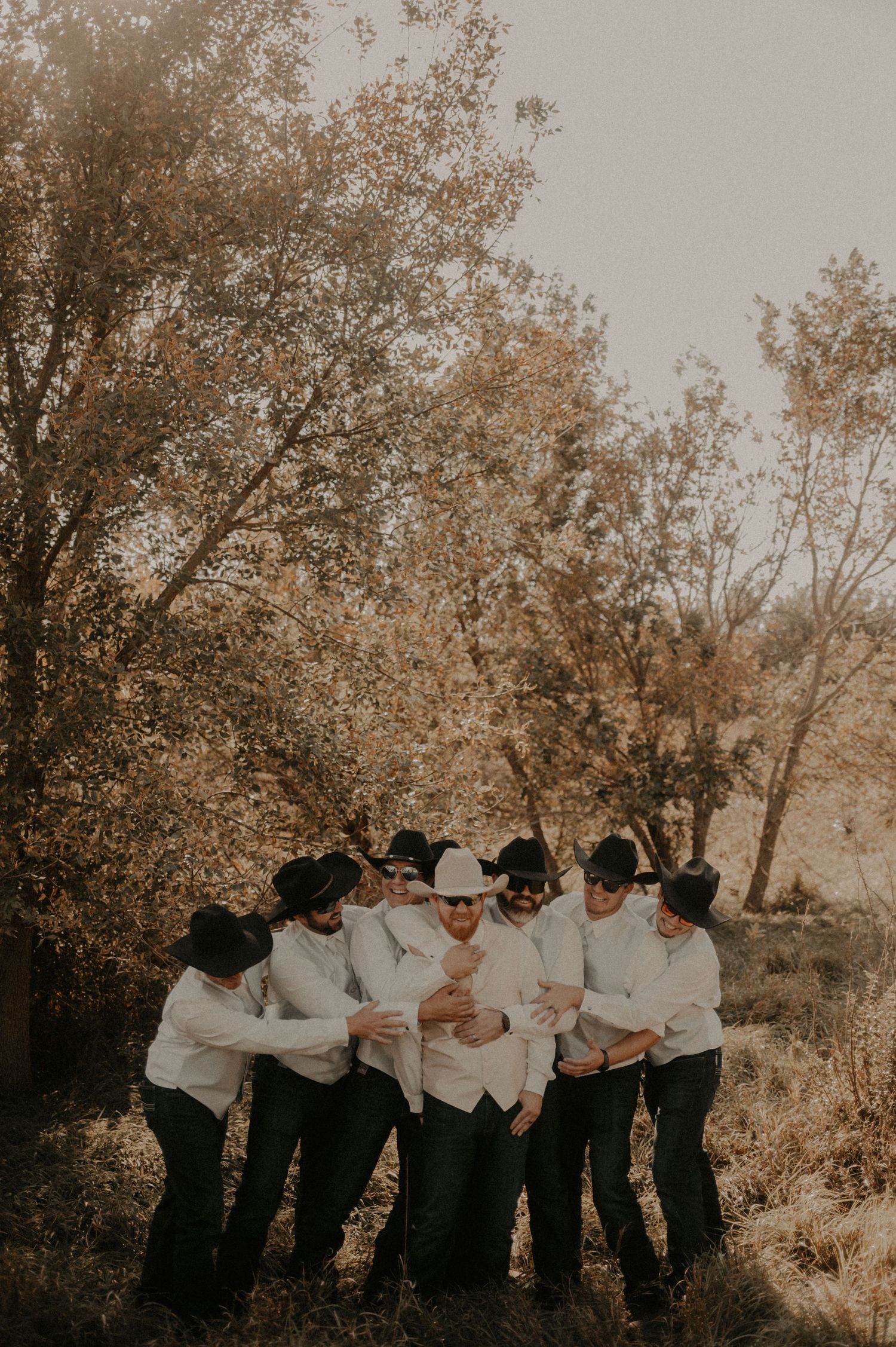 A group in matching white shirts and dark pants pose playfully together outdoors during a golden sunset.