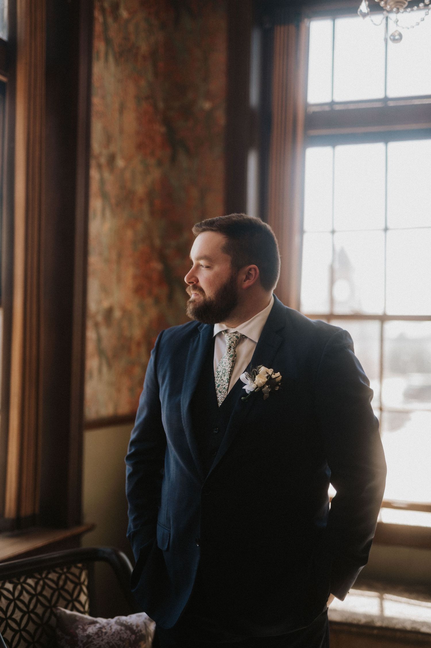 A groom in a navy suit gazes thoughtfully out of rustic windows in a warm-lit room during a wedding.