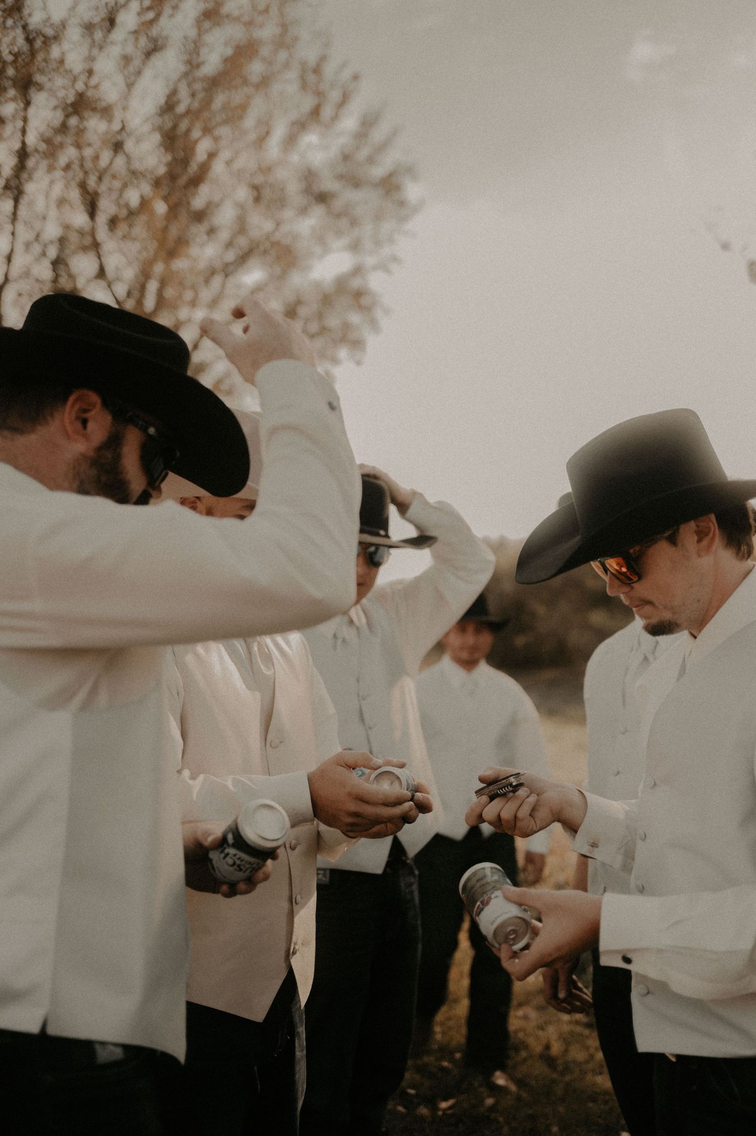 Groomsmen in white shirts and cowboy hats sharing drinks together outdoors.
