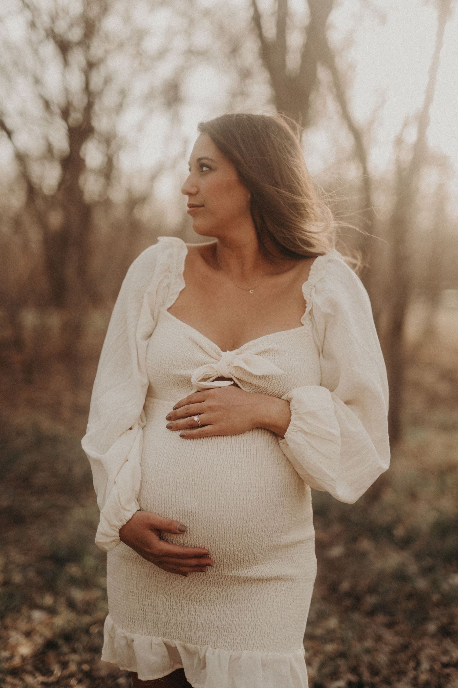 Woman in white puffy sleeve dress holds her pregnant belly in a romantic outdoor setting.