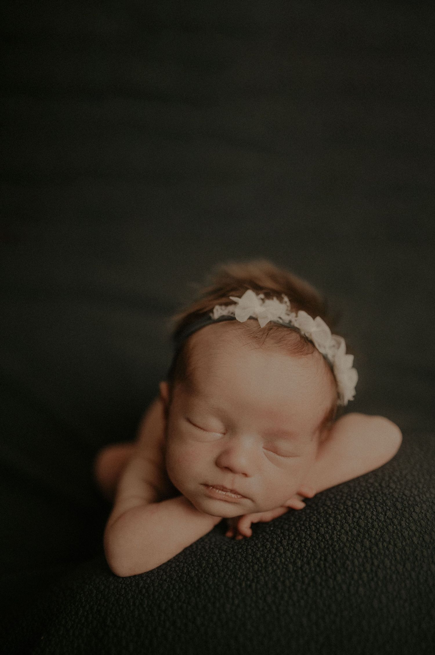 Minimalist newborn photography against dark background with delicate headpiece.
