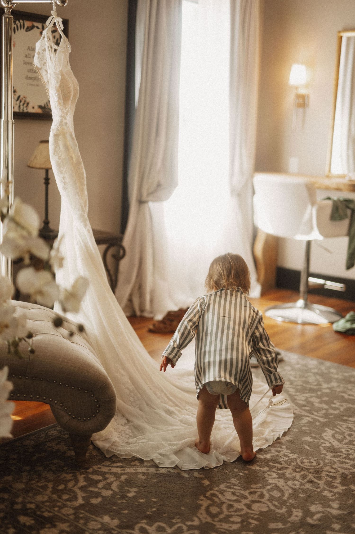 A flowing white wedding dress train trails across a patterned carpet in a sunlit room.