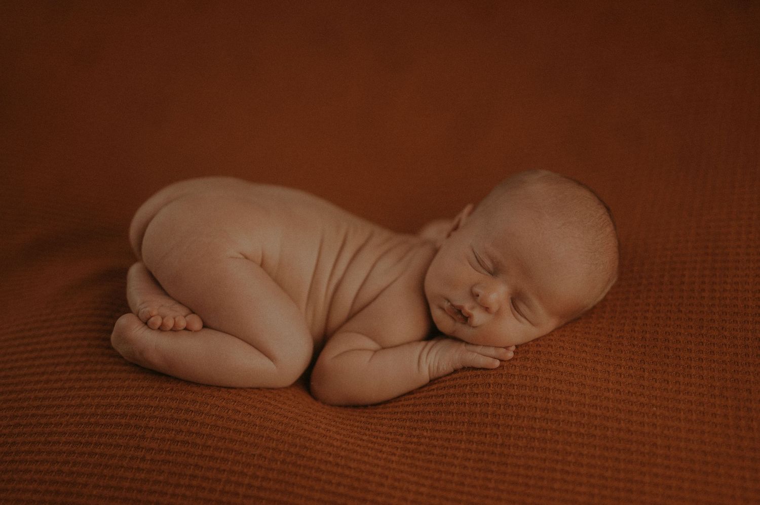 Peaceful sleeping newborn baby curled up on brown textured fabric in natural lighting.