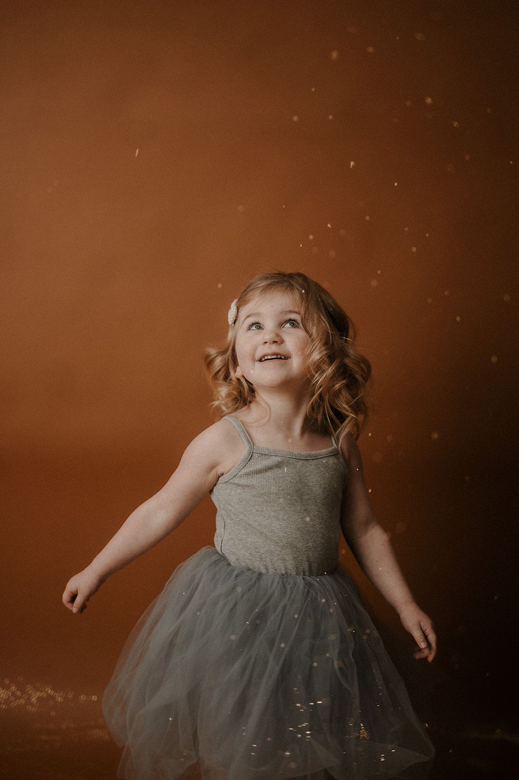 A child in a gray tulle dress spins and plays joyfully against a moody brown backdrop.