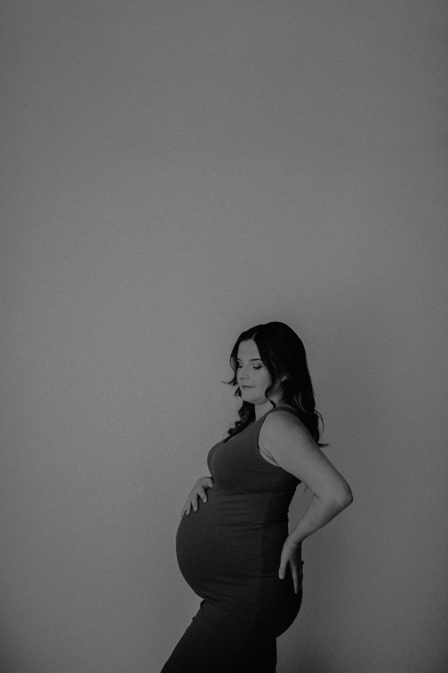 Black and white maternity portrait showing an expectant mother in a dark dress posing against a plain wall.