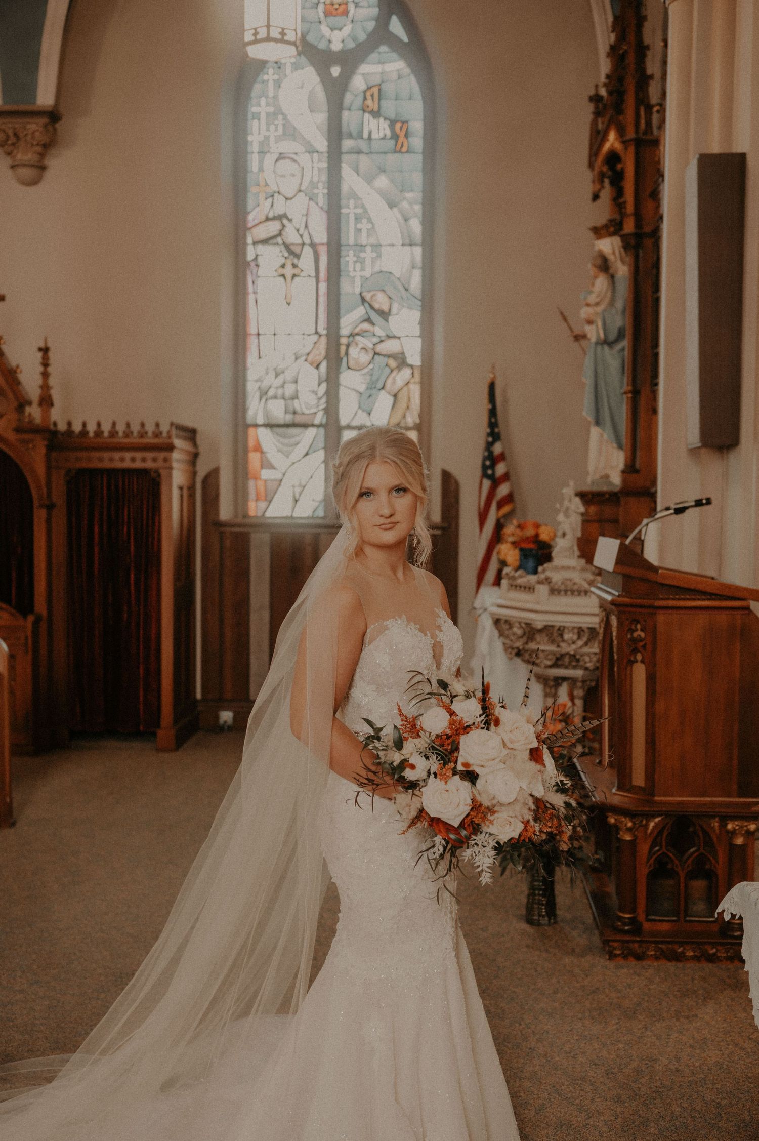 A bride in a flowing white gown and veil stands in a church interior with stained glass windows and wooden details.