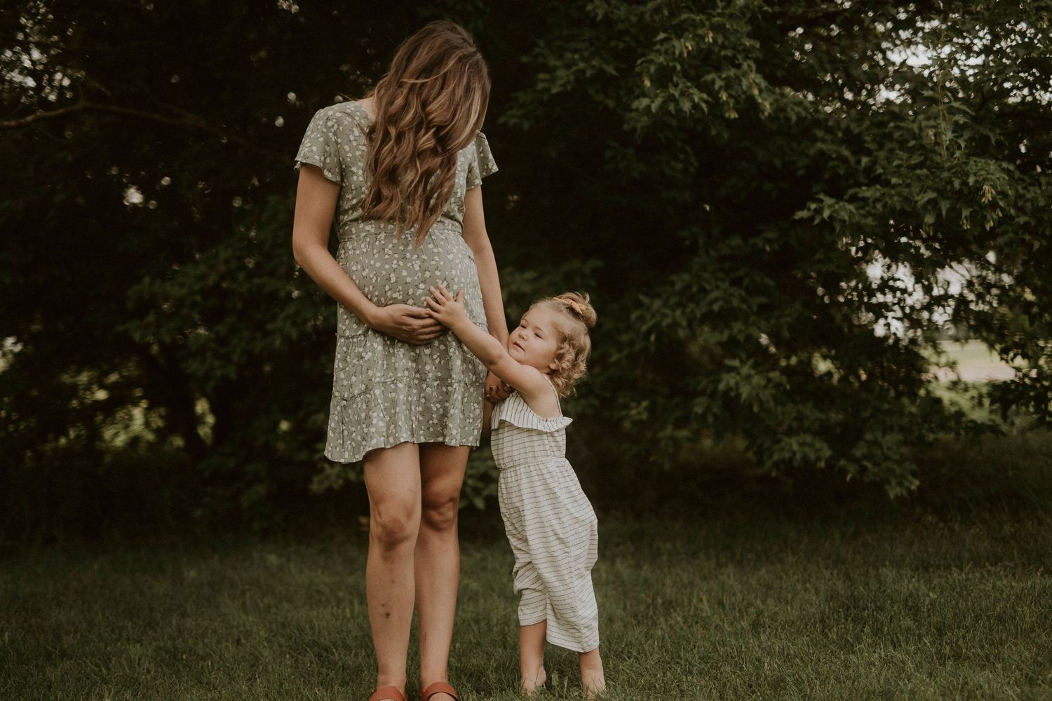 A mother and child walk together through a lush green garden setting in warm summer light.