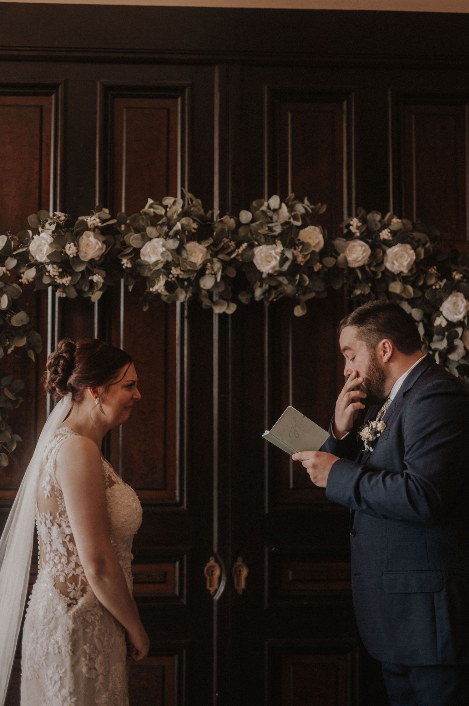 A couple exchanges vows beneath an elegant floral arch against dark wooden doors.