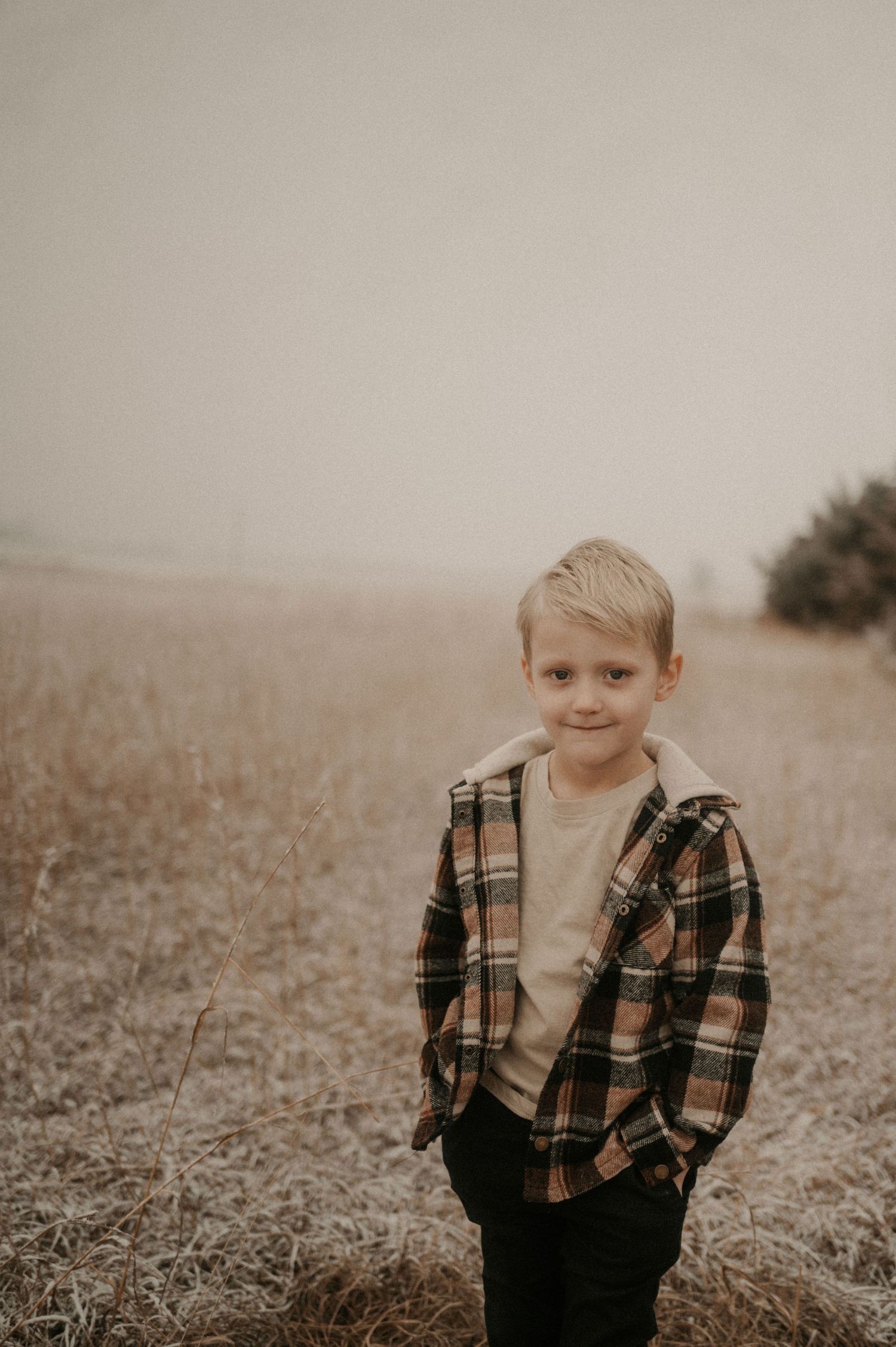 A child wearing a plaid jacket stands in a misty field with a soft sepia-toned aesthetic.