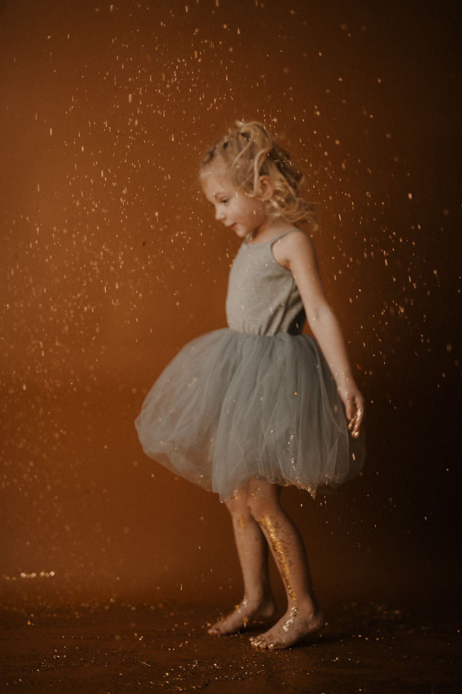 Young child in a grey tulle dress twirls and plays with glitter in a dreamy brown-toned setting.