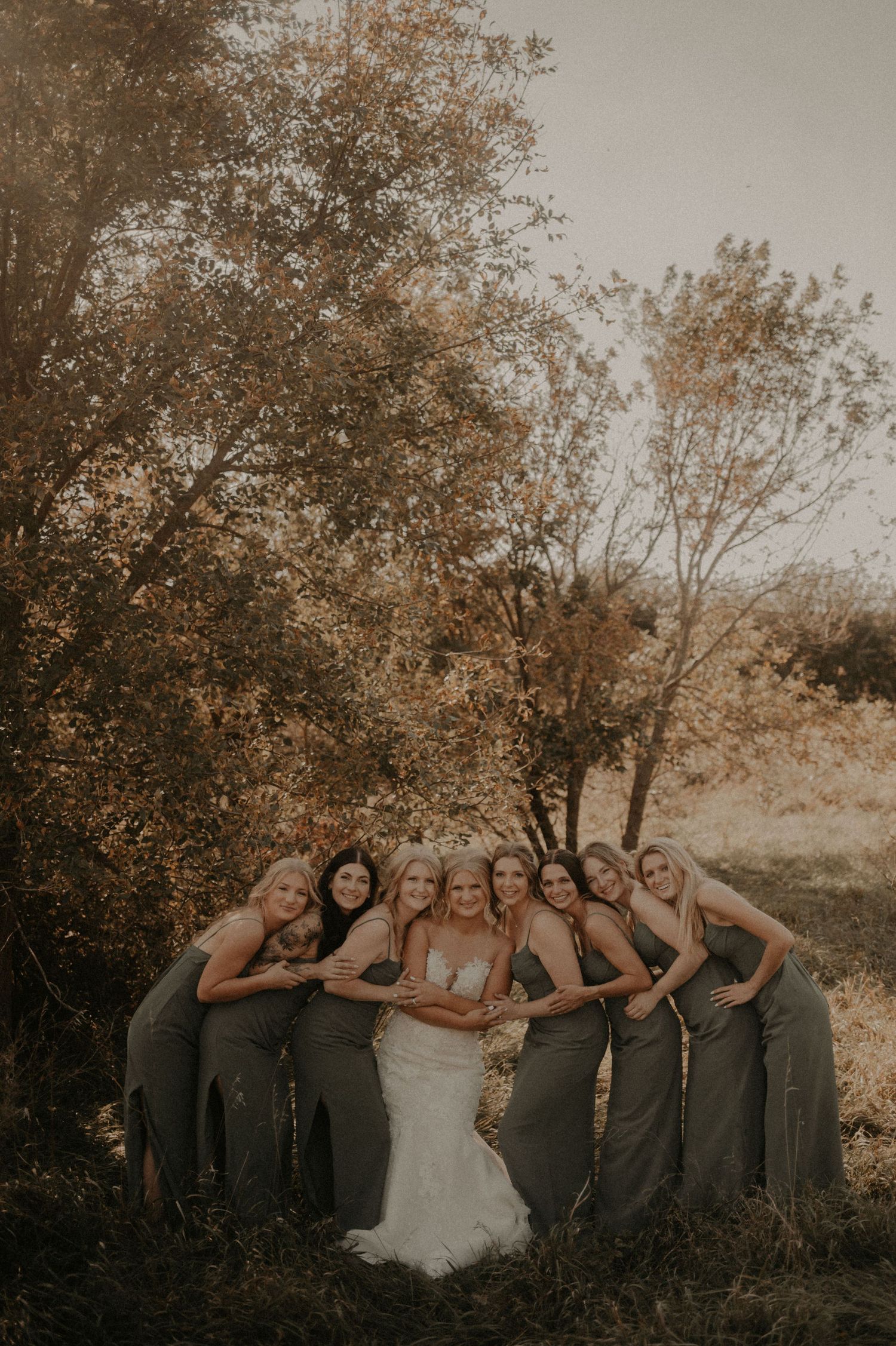 Group photo of bridesmaids in matching dresses posed together outdoors against autumn foliage.