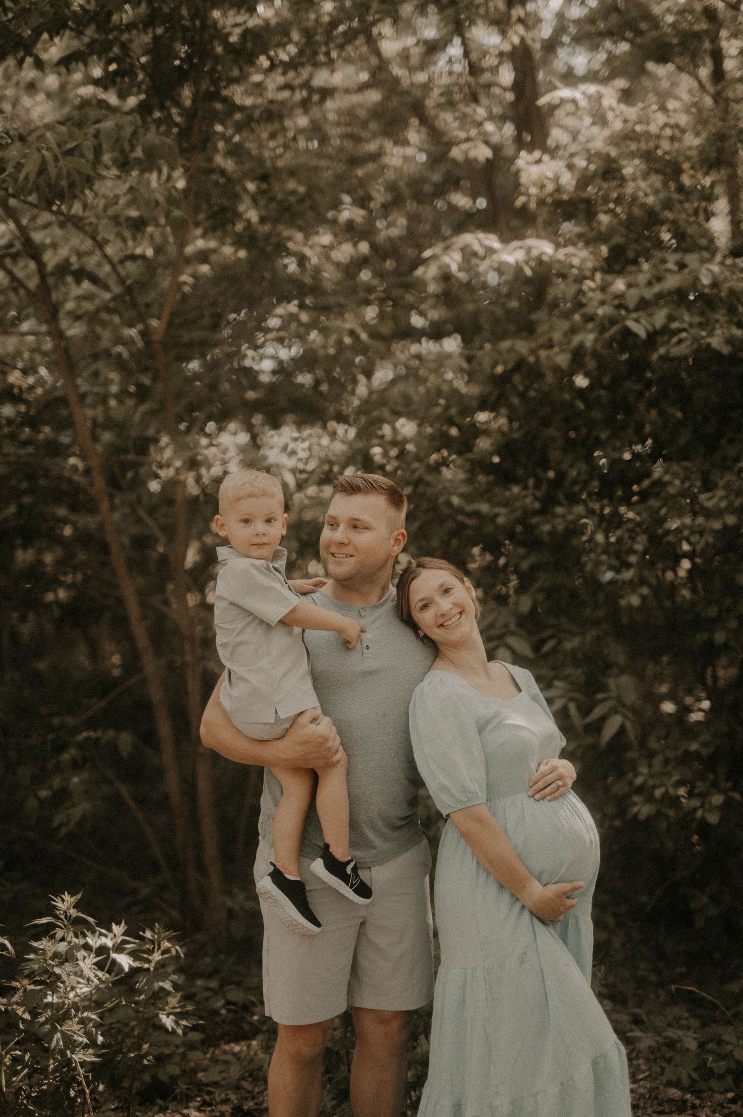 Family in coordinating gray and blue outfits pose together in a wooded outdoor setting during golden hour.