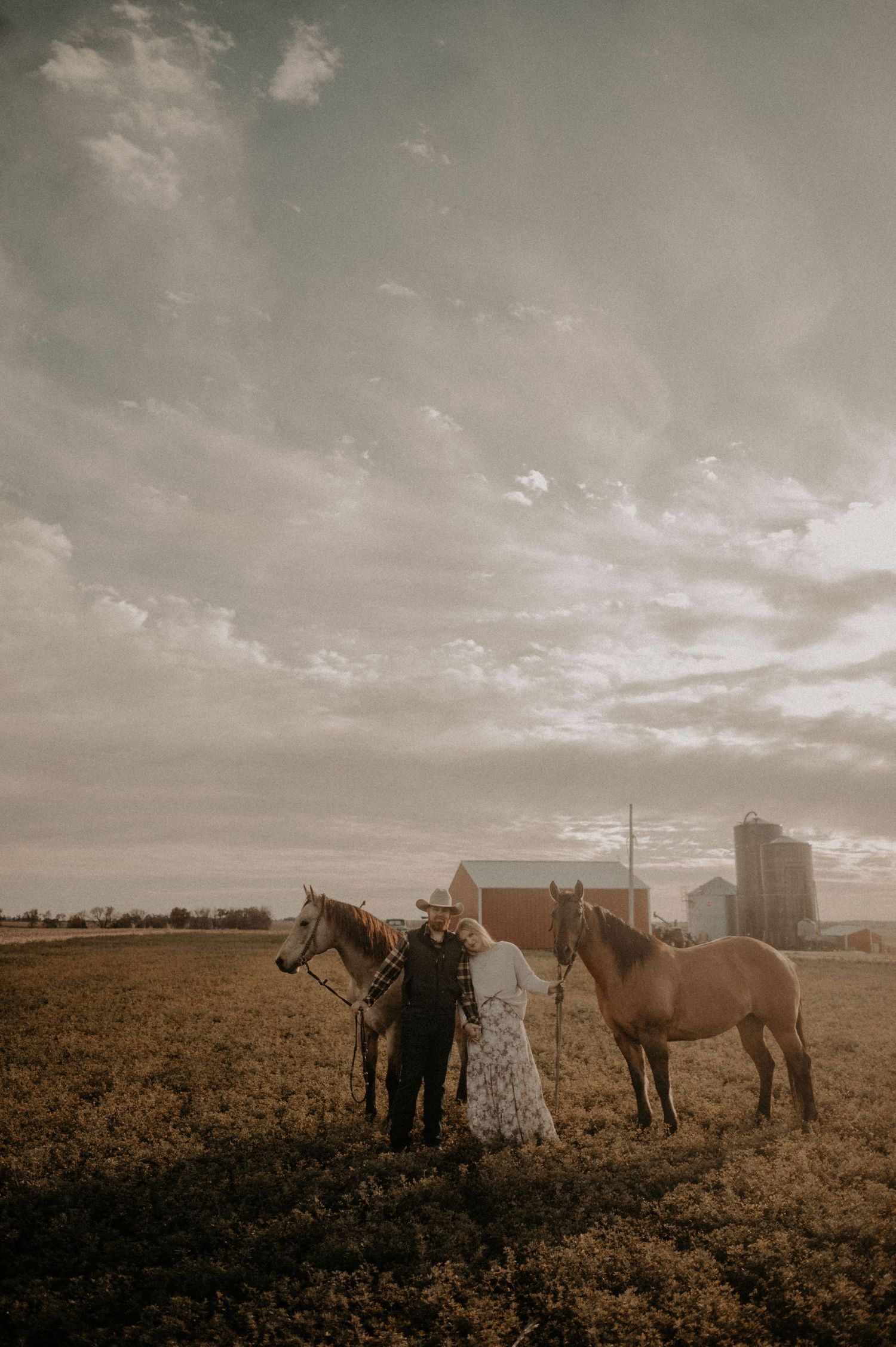 A rustic farm scene at sunset with two horses and people standing in a field with a barn and silo in the background.