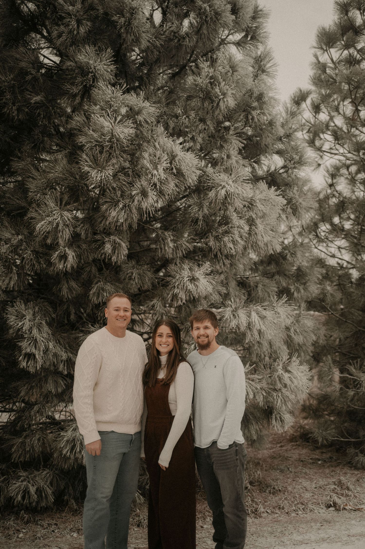 Three people pose together in a casual outdoor setting with pine trees in the background in black and white.