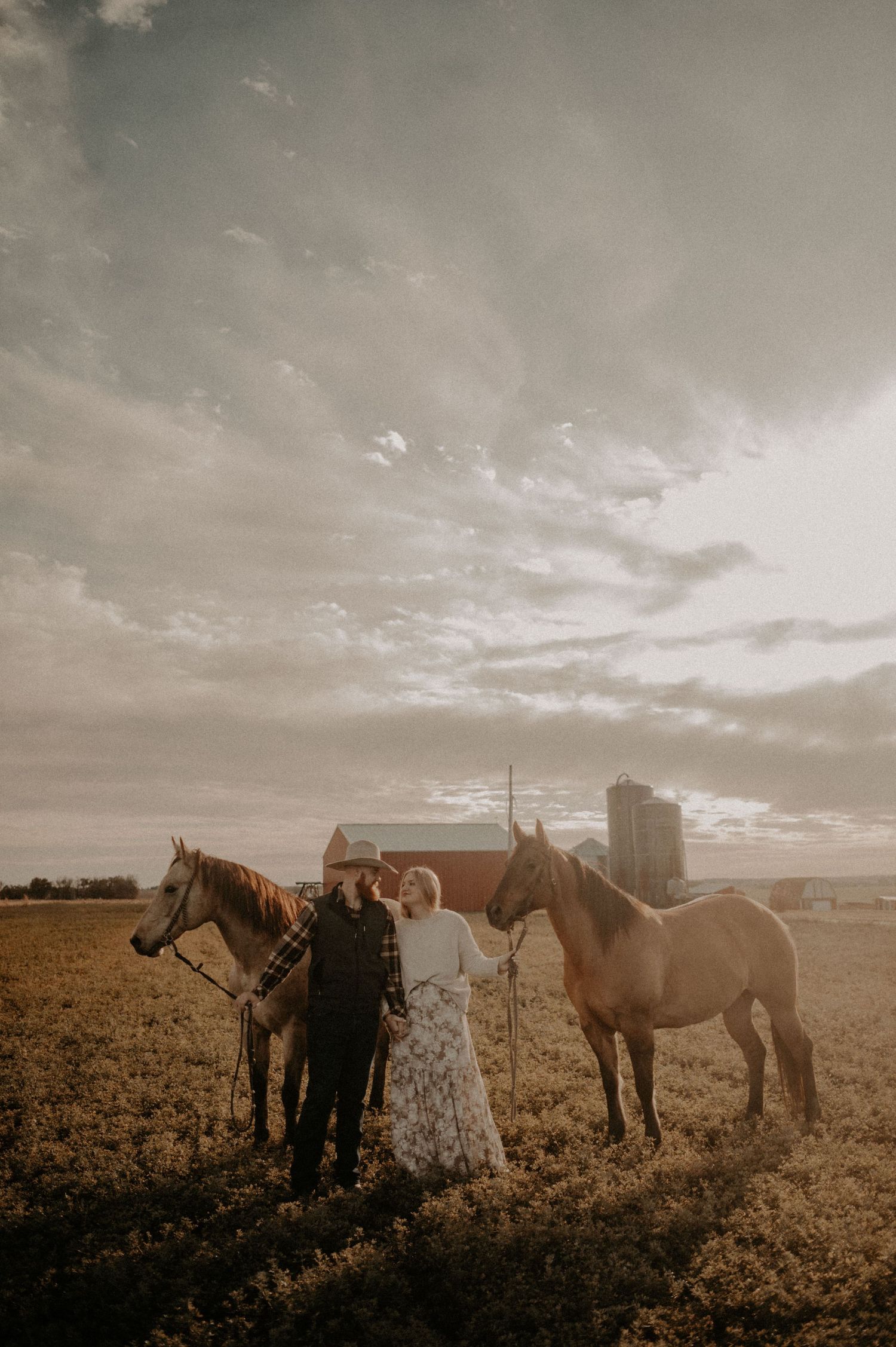 A wedding couple and their horses pose in a field during sunset with city buildings in background.