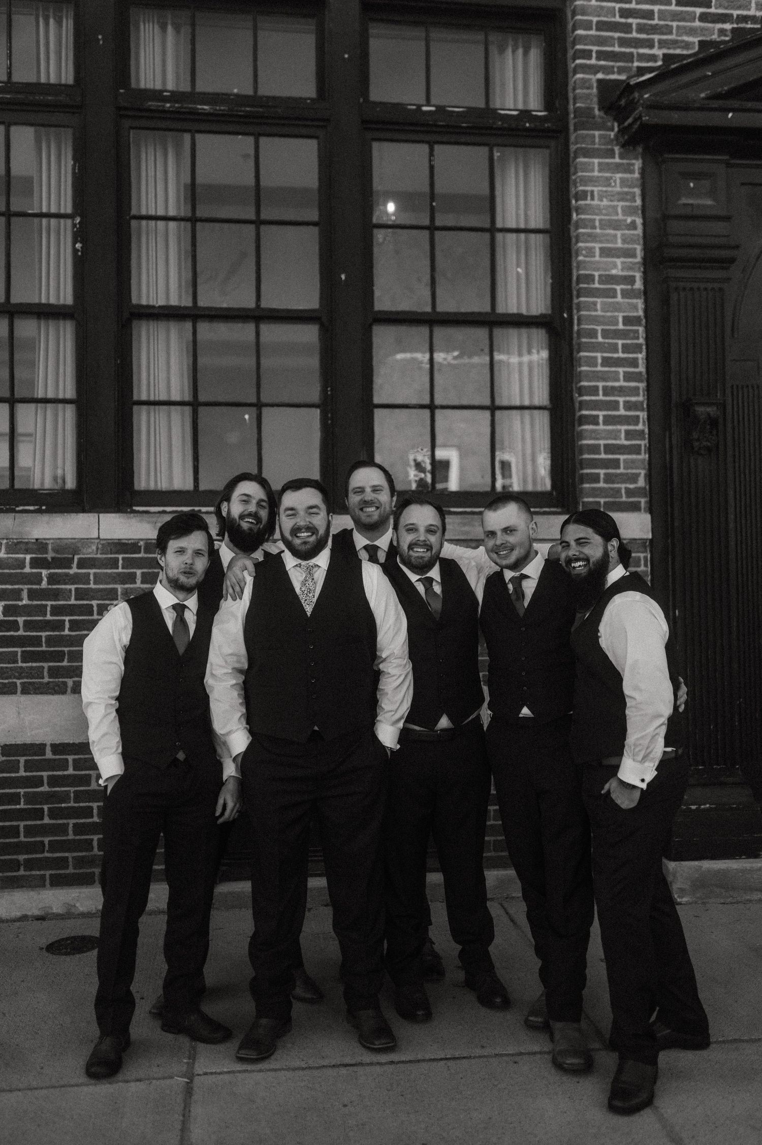 Groomsmen in vests and suits pose casually outside brick building.