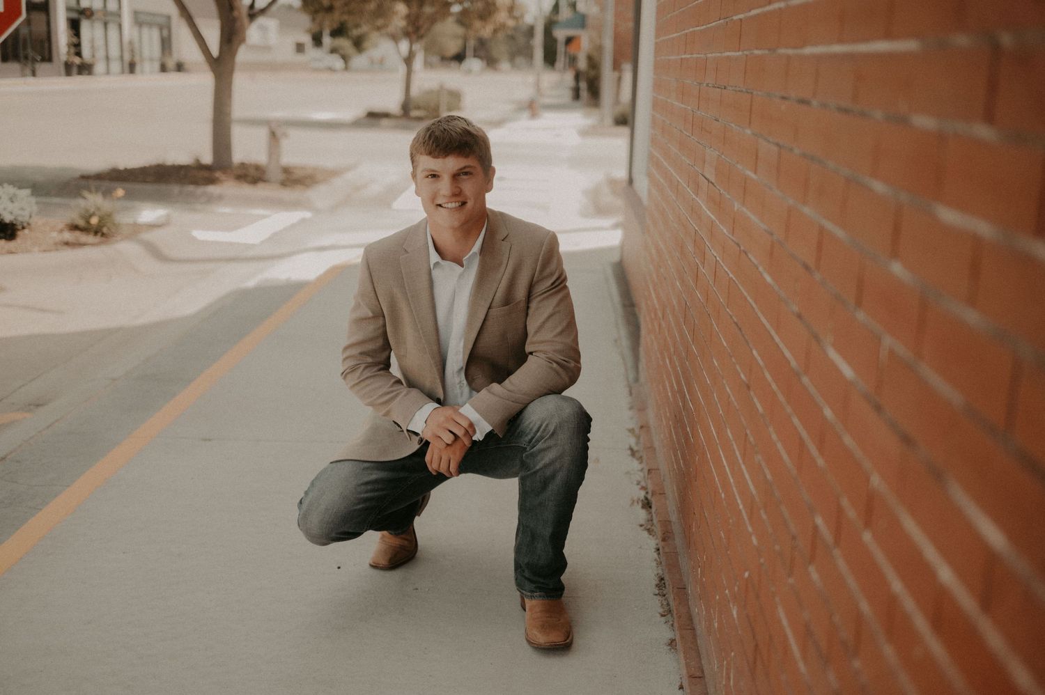 A person in a brown blazer and gray pants sits casually against a brick wall in an outdoor setting.