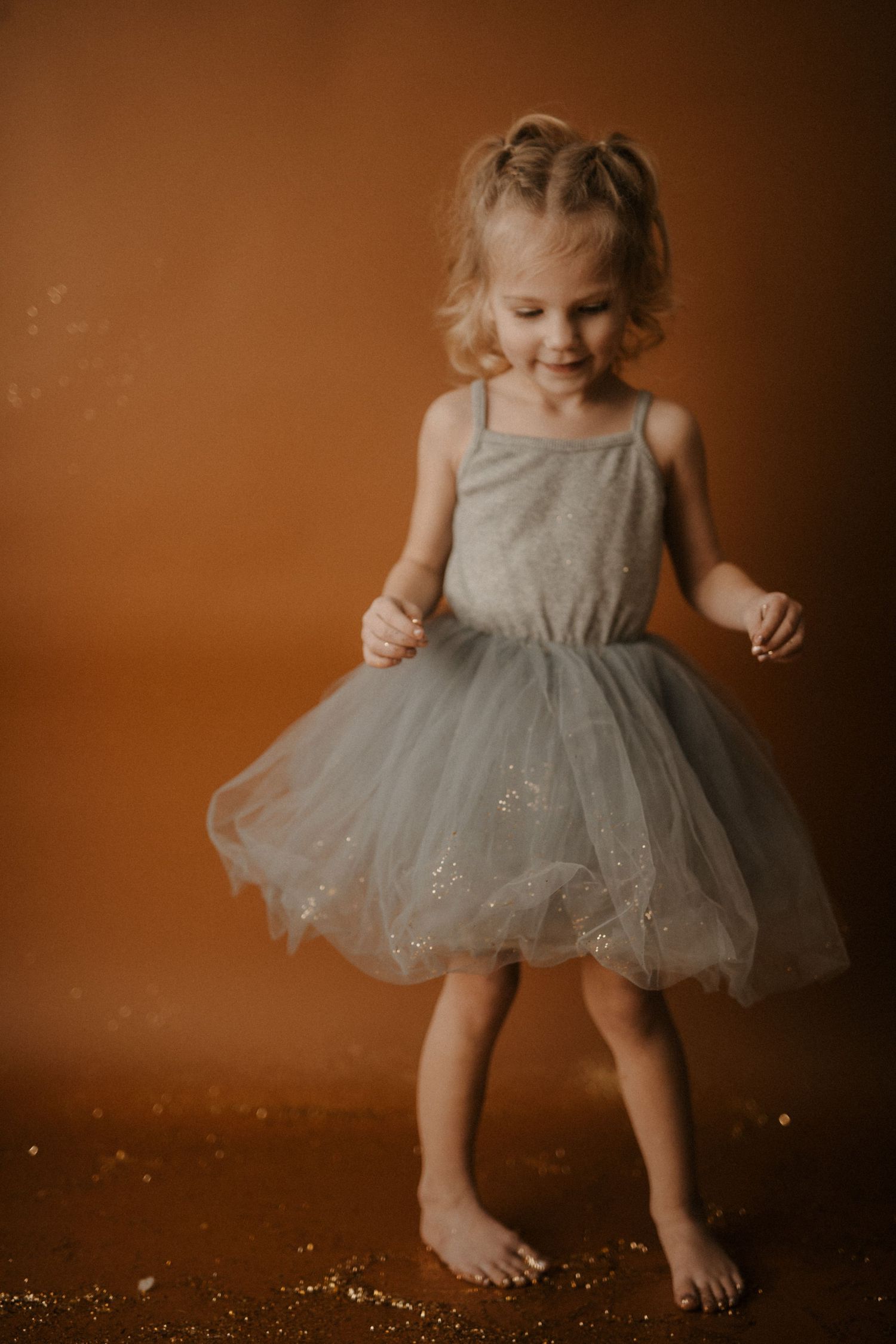 A toddler in a sparkly gray tulle dress twirls and dances against a warm brown backdrop in a series of motion shots.