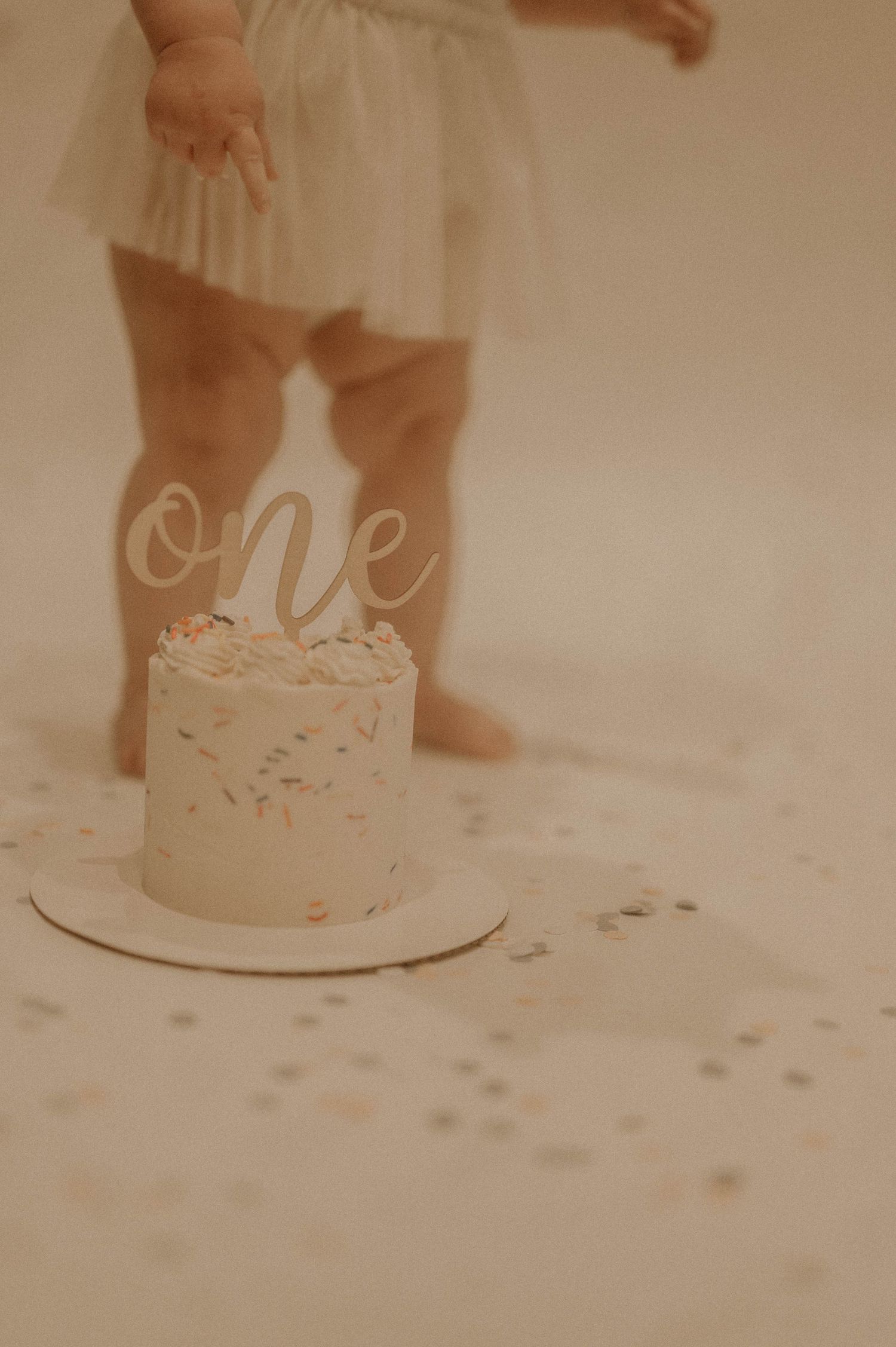 Small white birthday cake with 'One' topper and tiny feet visible in the background against a neutral backdrop.
