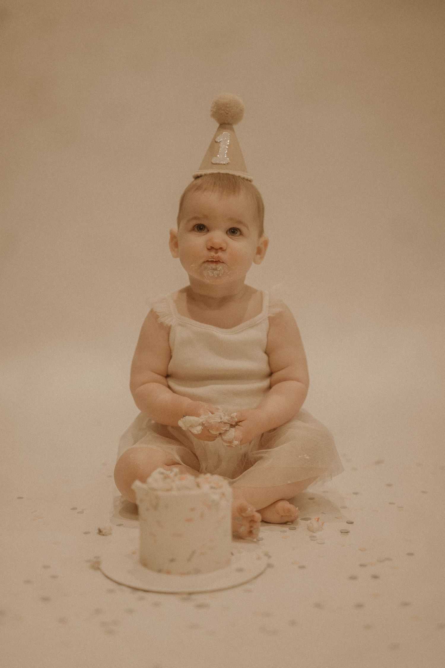 A baby in a white outfit sits on the floor with a birthday cake, showing progression of eating and making a mess.