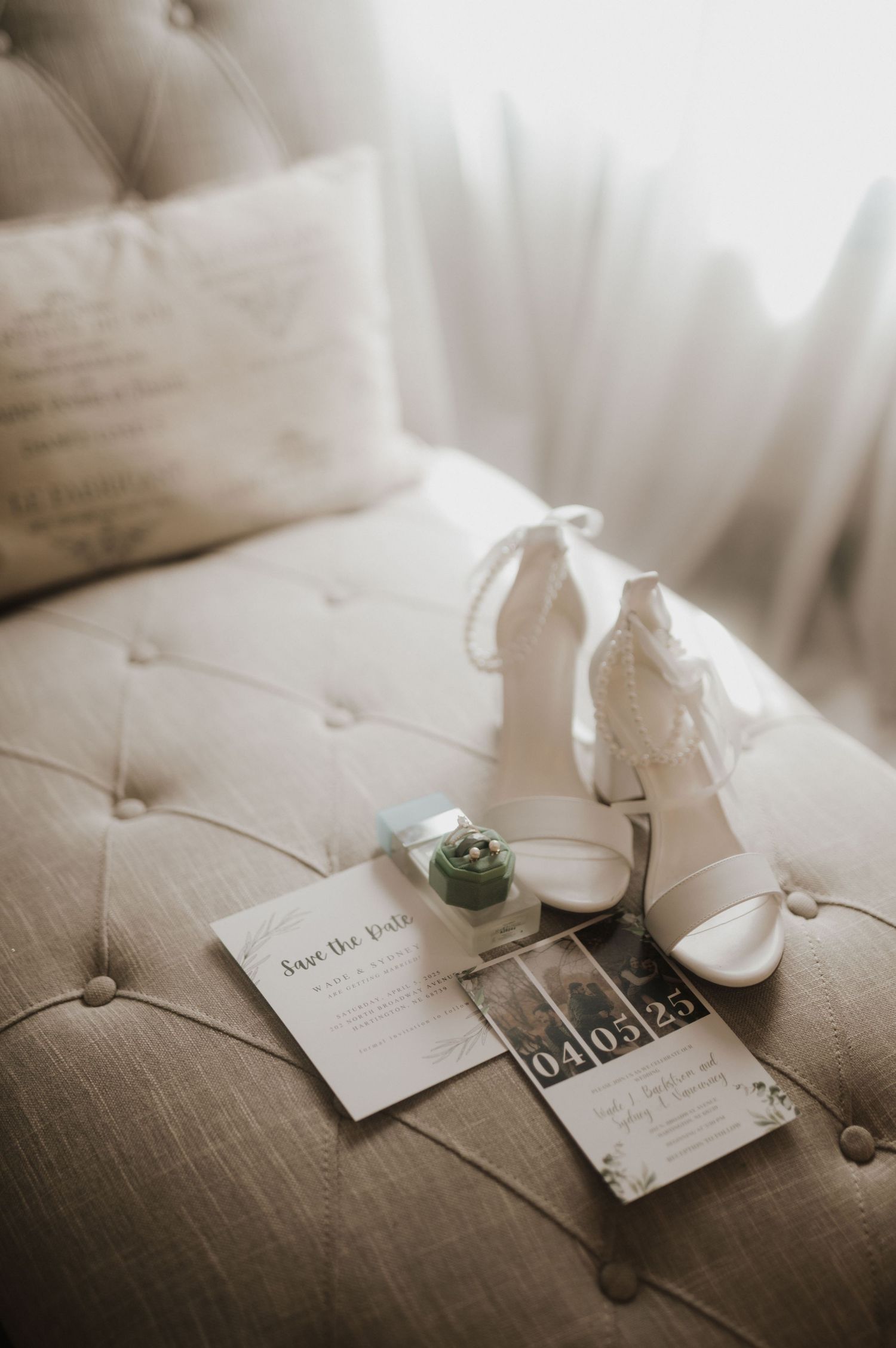 Wedding invitation cards and white heels arranged on beige tufted surface.