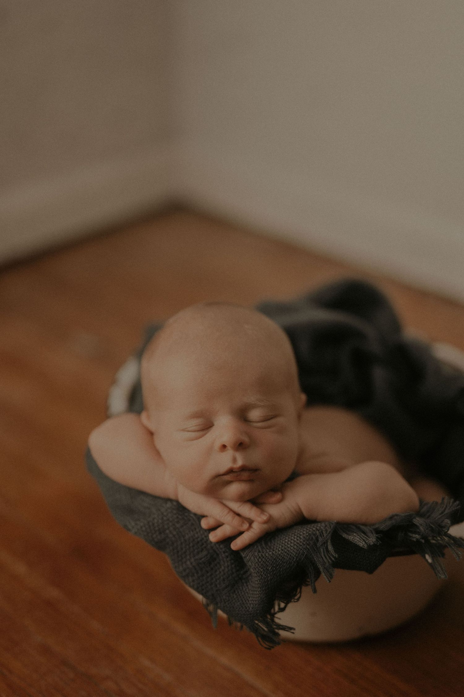 A sleeping newborn baby wrapped in a dark gray blanket rests peacefully on a wooden surface in warm lighting.