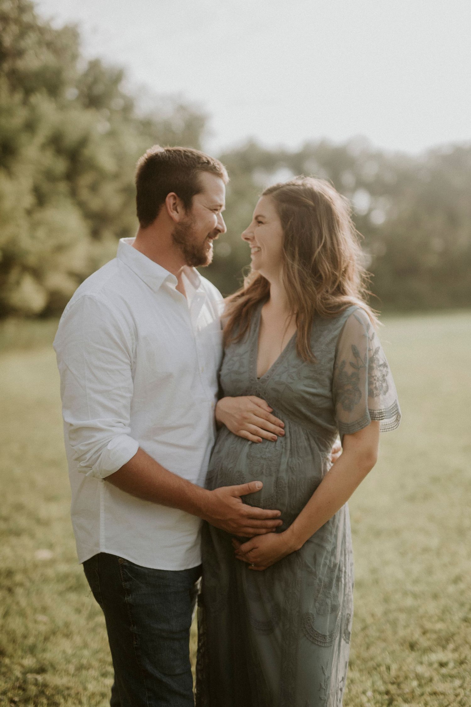 A couple shares an intimate moment outdoors as they lovingly embrace a pregnant belly in soft sunlit scenery.