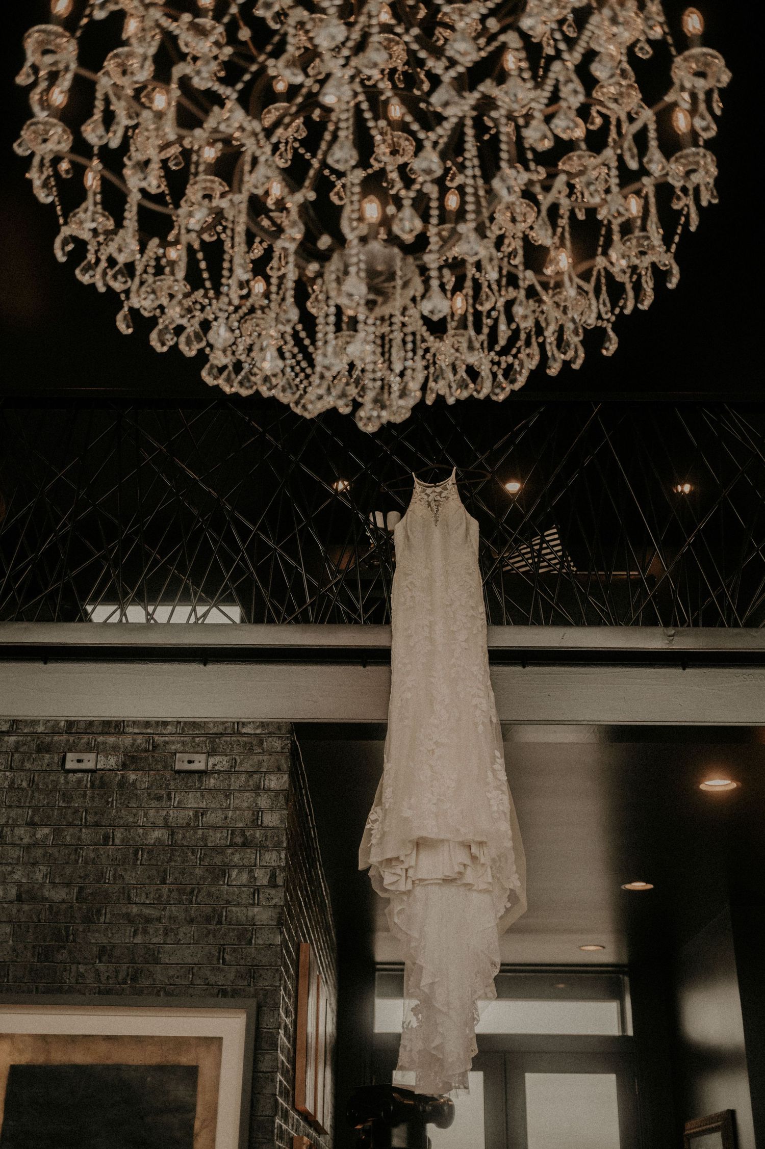 An elegant white wedding dress hangs beneath a crystal chandelier in a rustic brick venue.