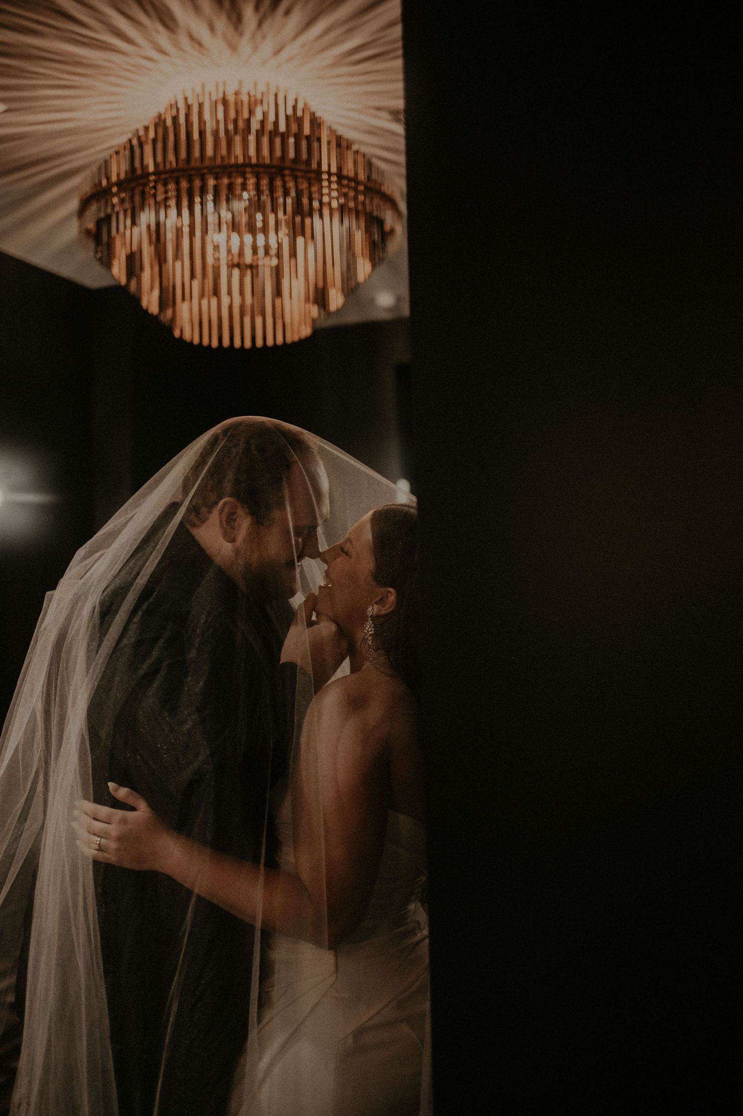 Elegant chandelier creates dramatic lighting above intimate wedding moment in dark reception venue.