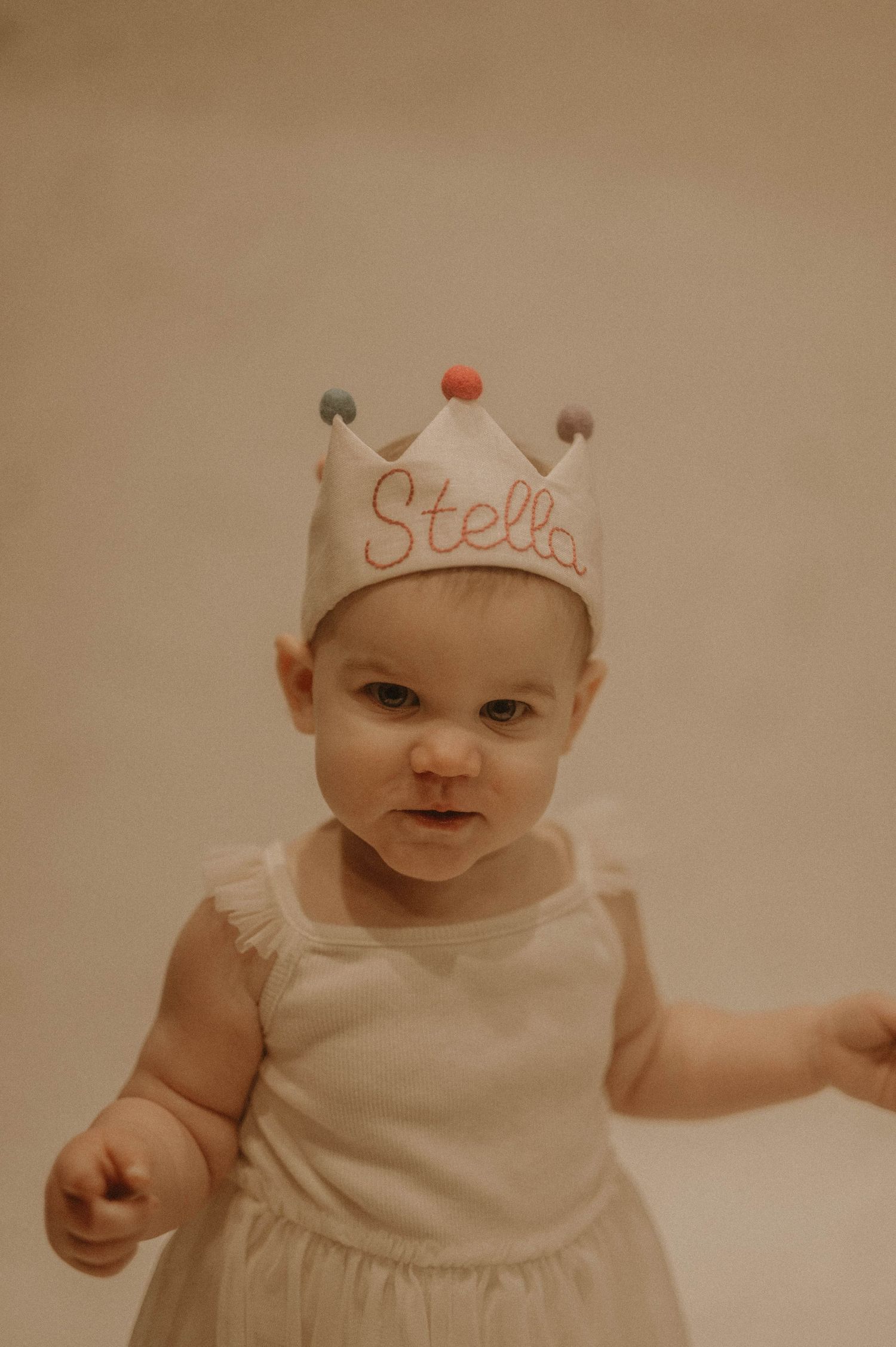 A small child wearing a white crown with the name 'Stella' and a white sleeveless dress against a neutral background.