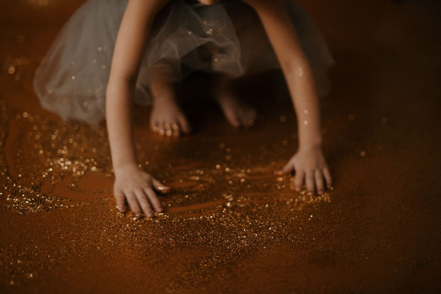Small hands touch glittery sparkles scattered across a dark wooden surface in moody lighting.