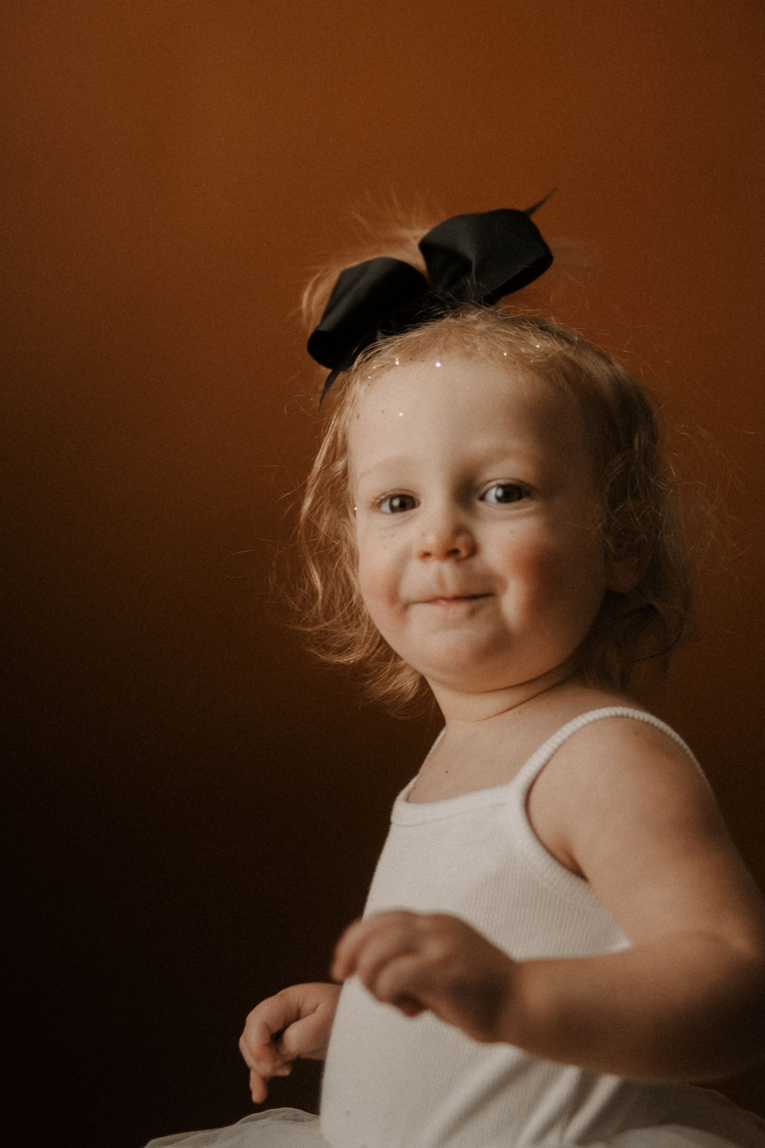A young child in a white tank top and black bow smiles warmly against a dark brown background.