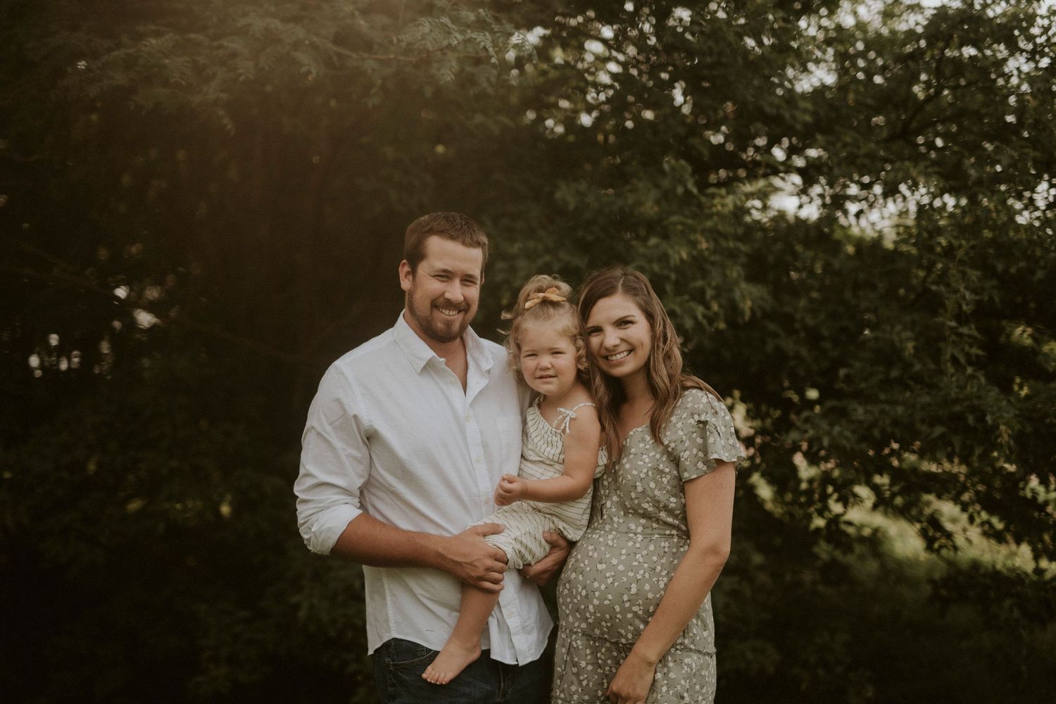 A family in white and neutral colored clothing embraces while standing in a grassy outdoor setting during golden hour.