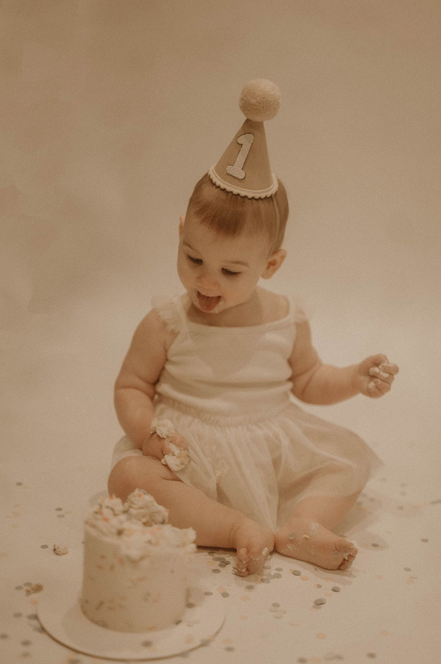 Baby wearing party hat and white dress sits on floor with small cake during first birthday photoshoot.
