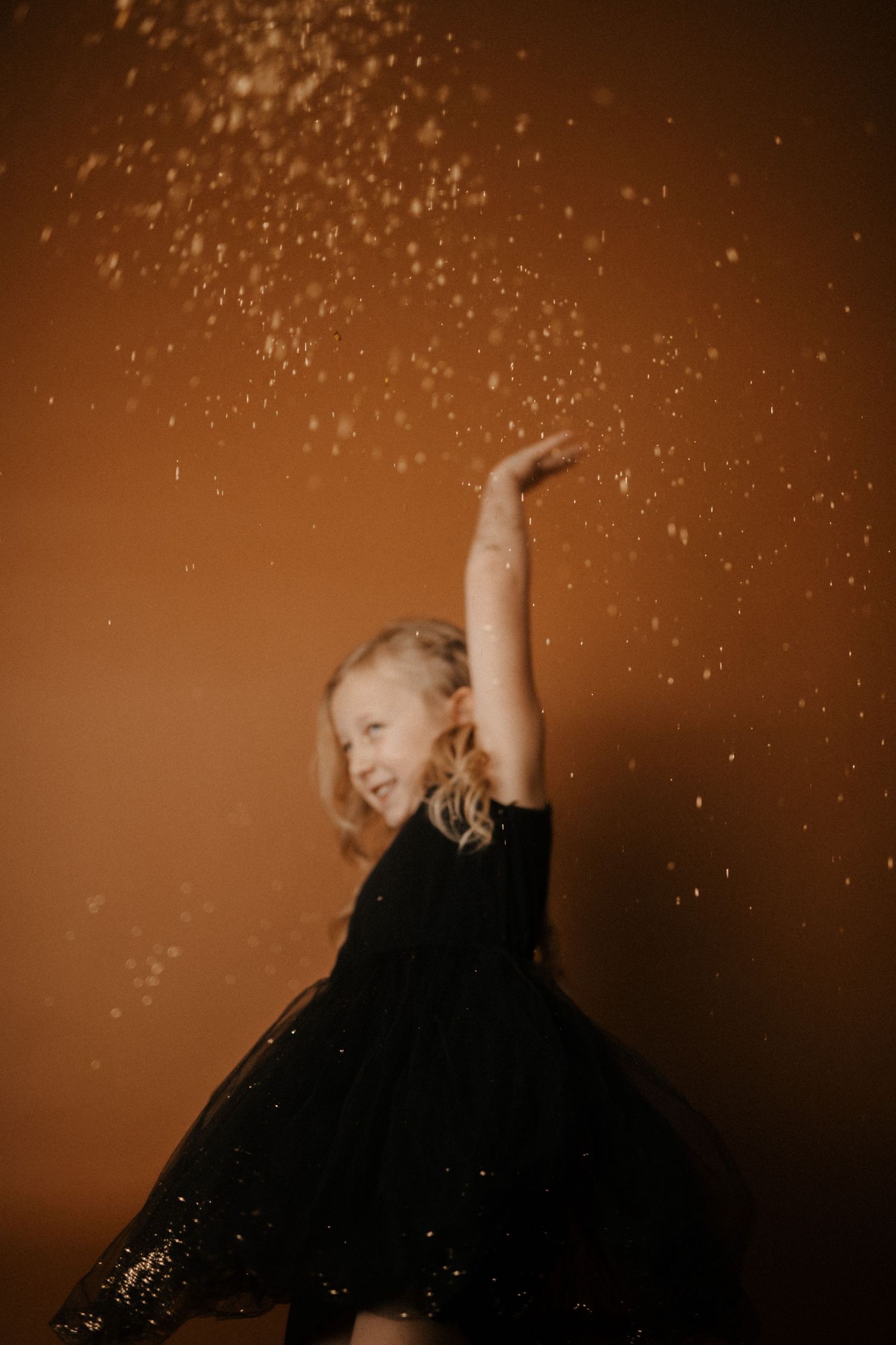 Figure in black dress dances expressively against warm brown background with sparkle effects in artistic portrait series.