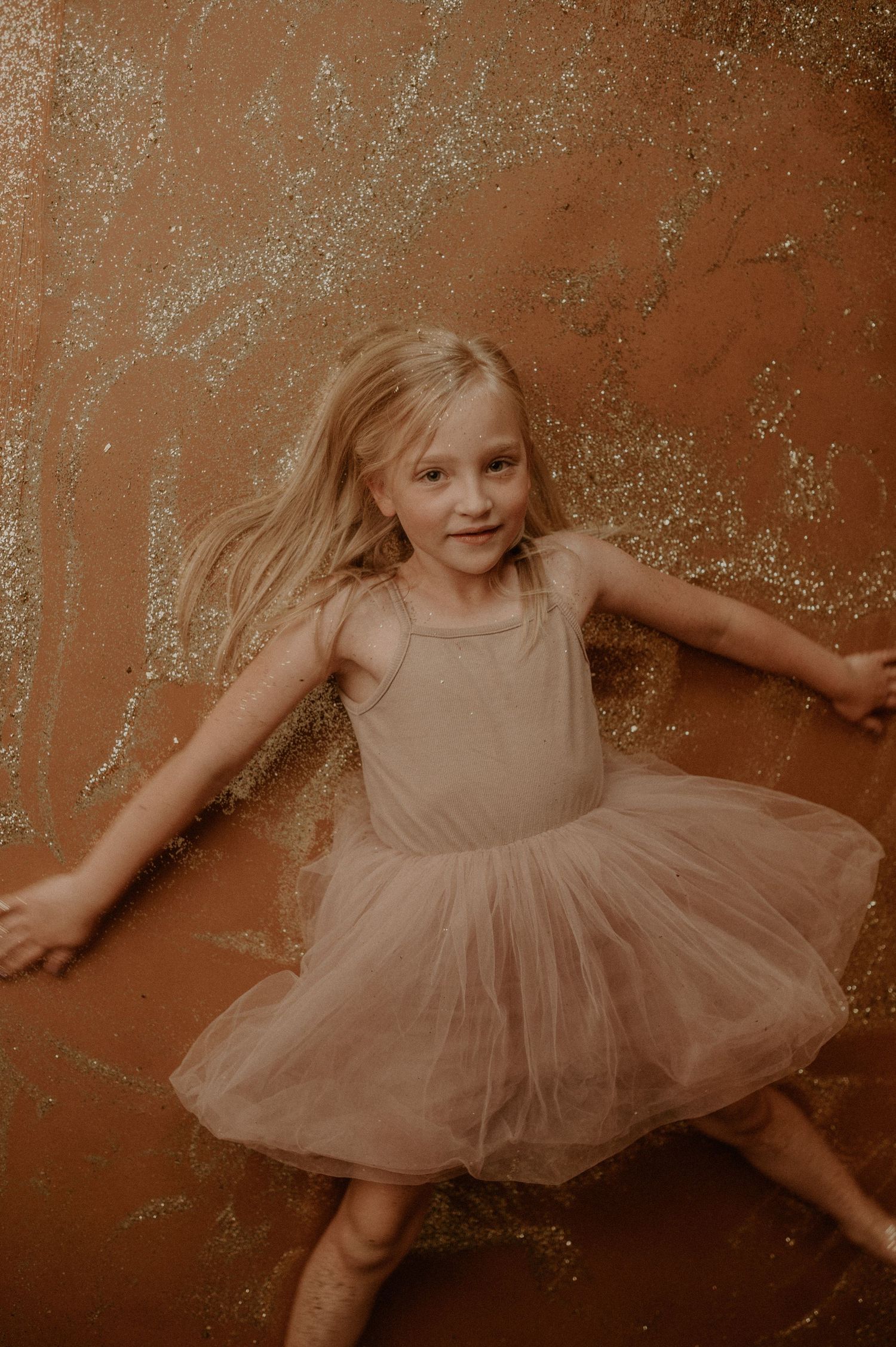 Young dancer in a beige tulle dress twirls joyfully against a rustic brown backdrop with sparkling details.