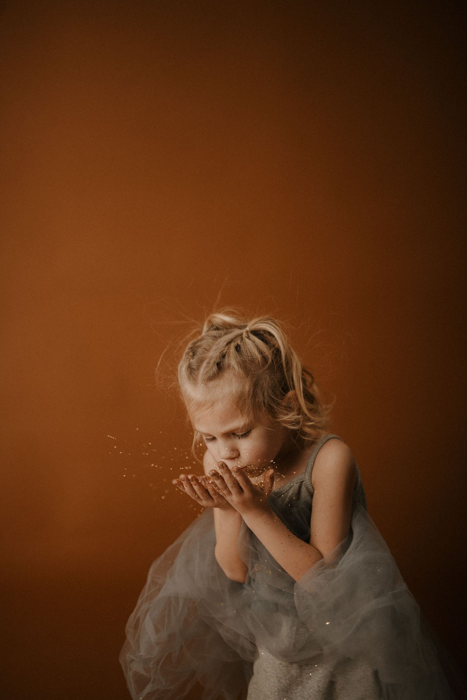 A child in a gray dress twirls and plays with flowing fabric in a warm, sepia-toned studio setting.