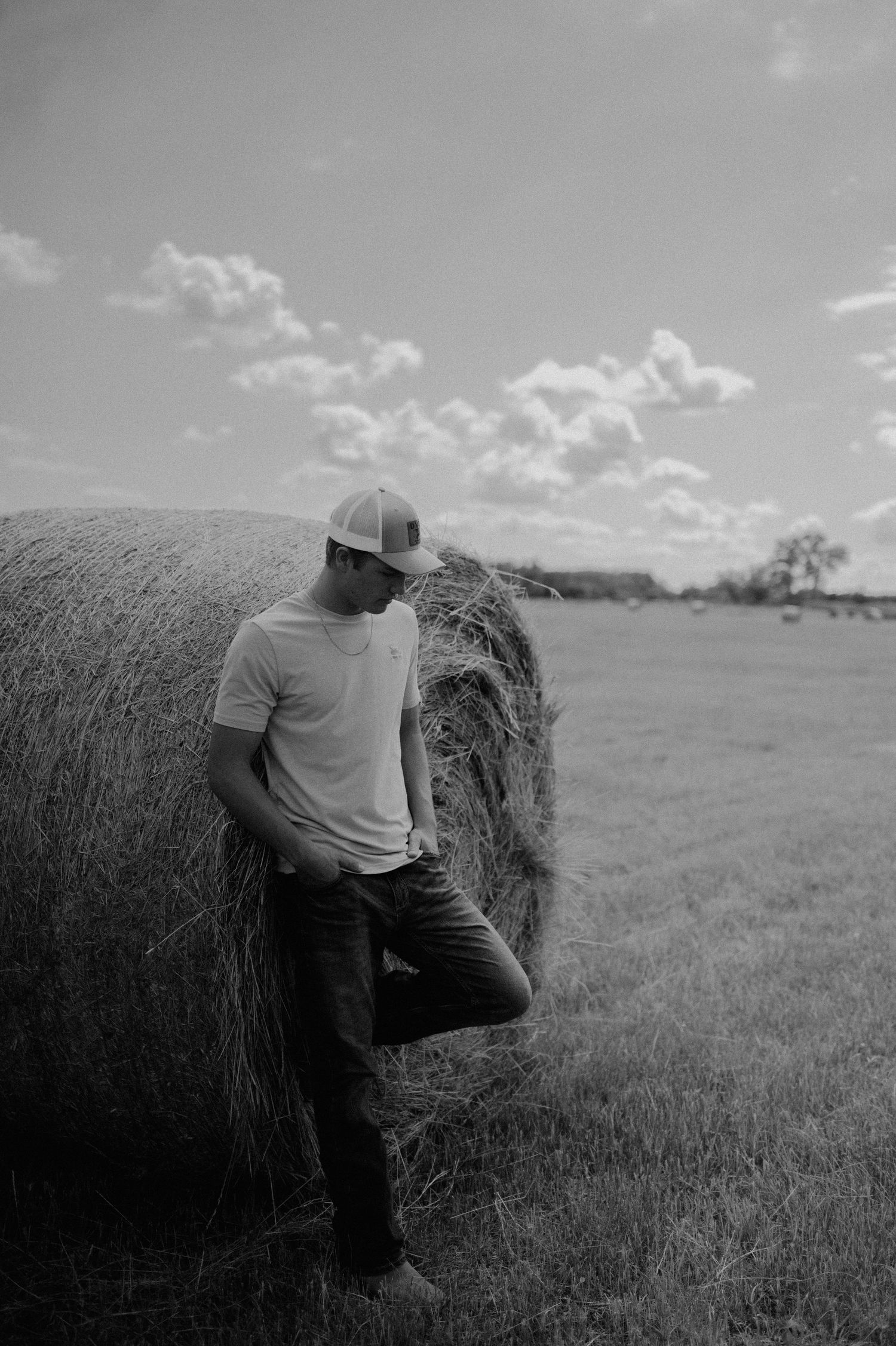 A person in a gray shirt demonstrates climbing technique on a hay bale in a rural field under cloudy skies.