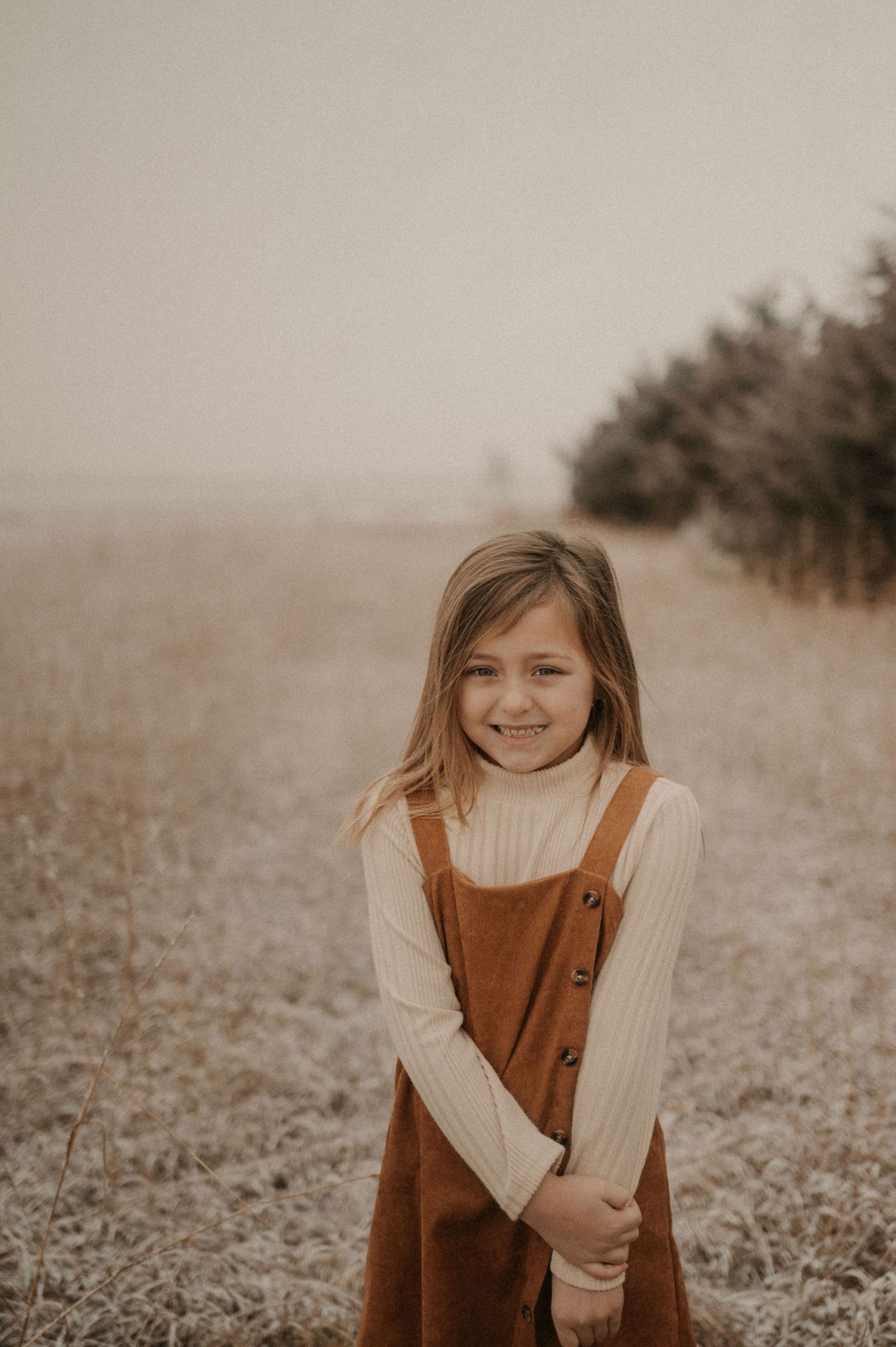 A child in a brown corduroy dress and cream sweater stands in a misty autumn field with trees in the background.