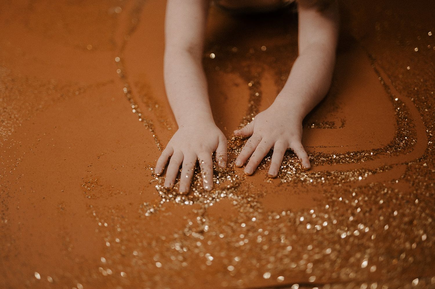 Small hands touching scattered glitter and sparkles on a rich brown background with dramatic lighting.