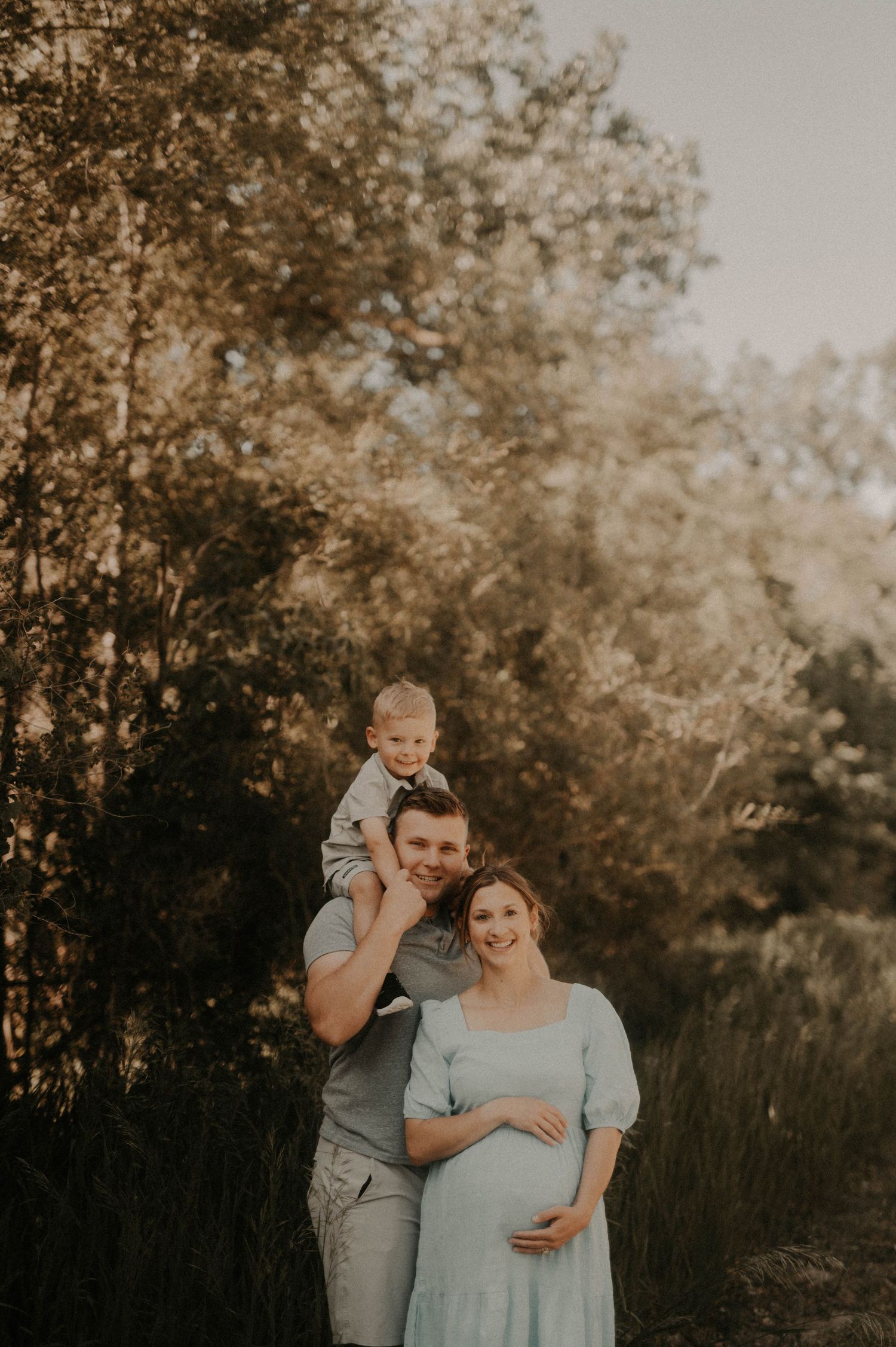 Family maternity portrait in nature showing couple with toddler on shoulders against warm sunset backdrop.