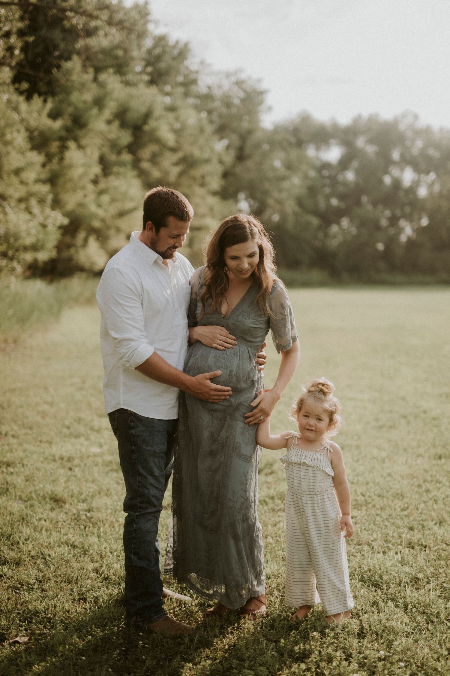 A family stands together in a sunlit field, with a pregnant mother in a gray dress and young child in white.