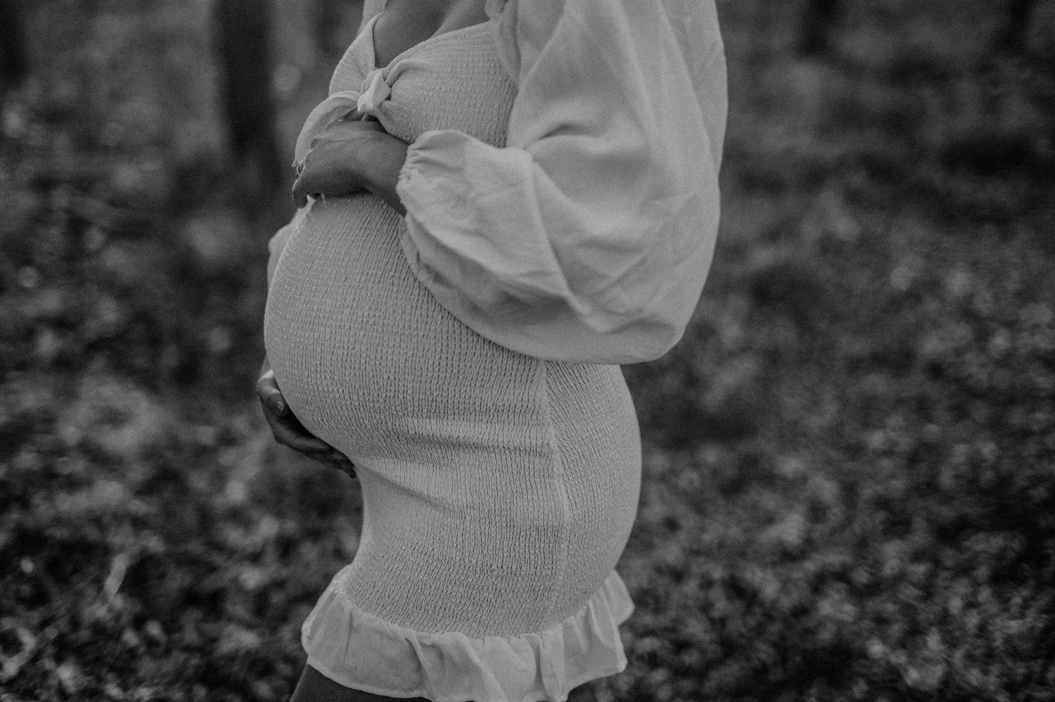 A dreamy maternity photo shows a woman in a white dress cradling her belly in soft sunlight.